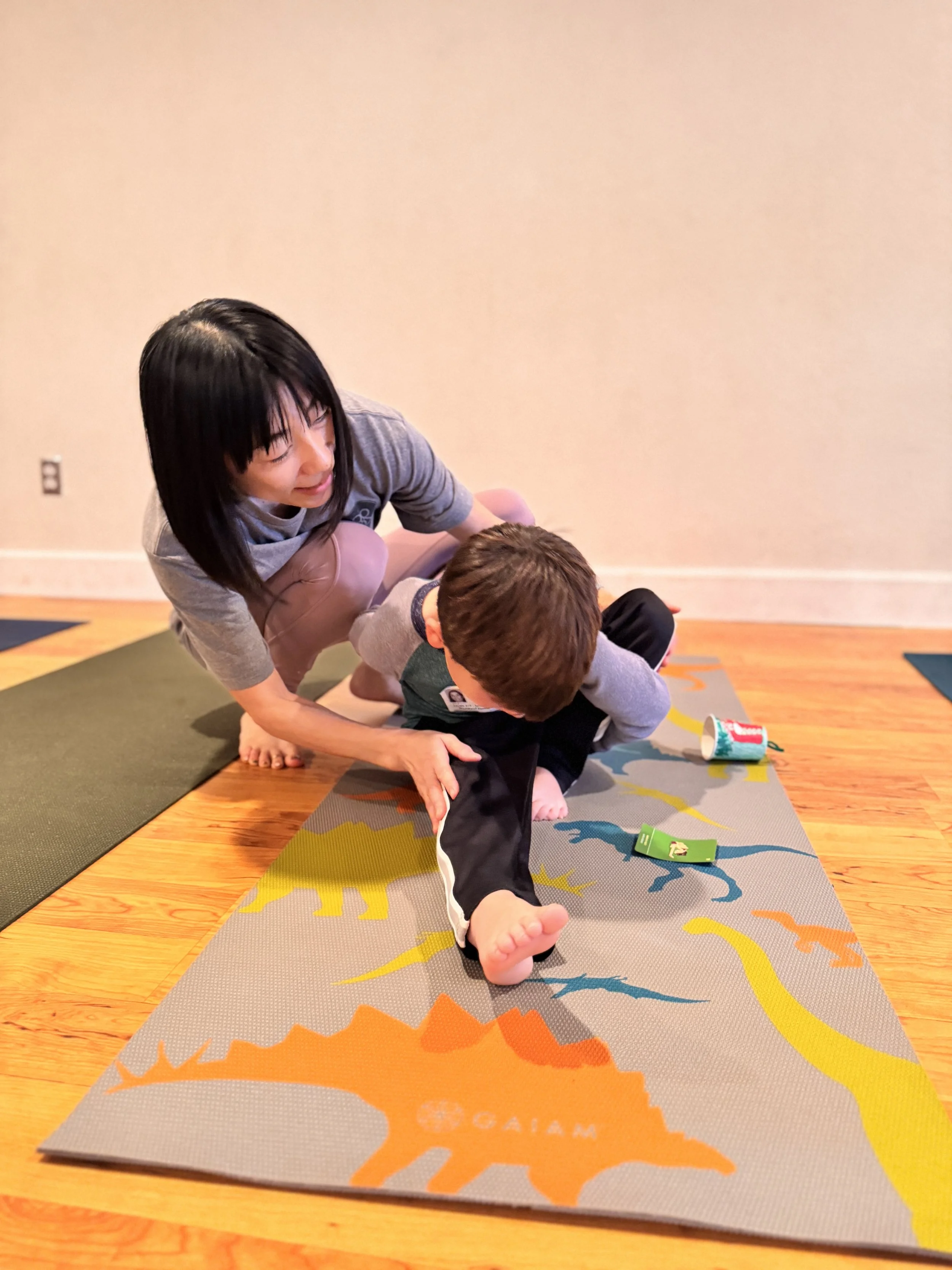 A woman assisting a young boy during a yoga pose on a colorful mat indoors.