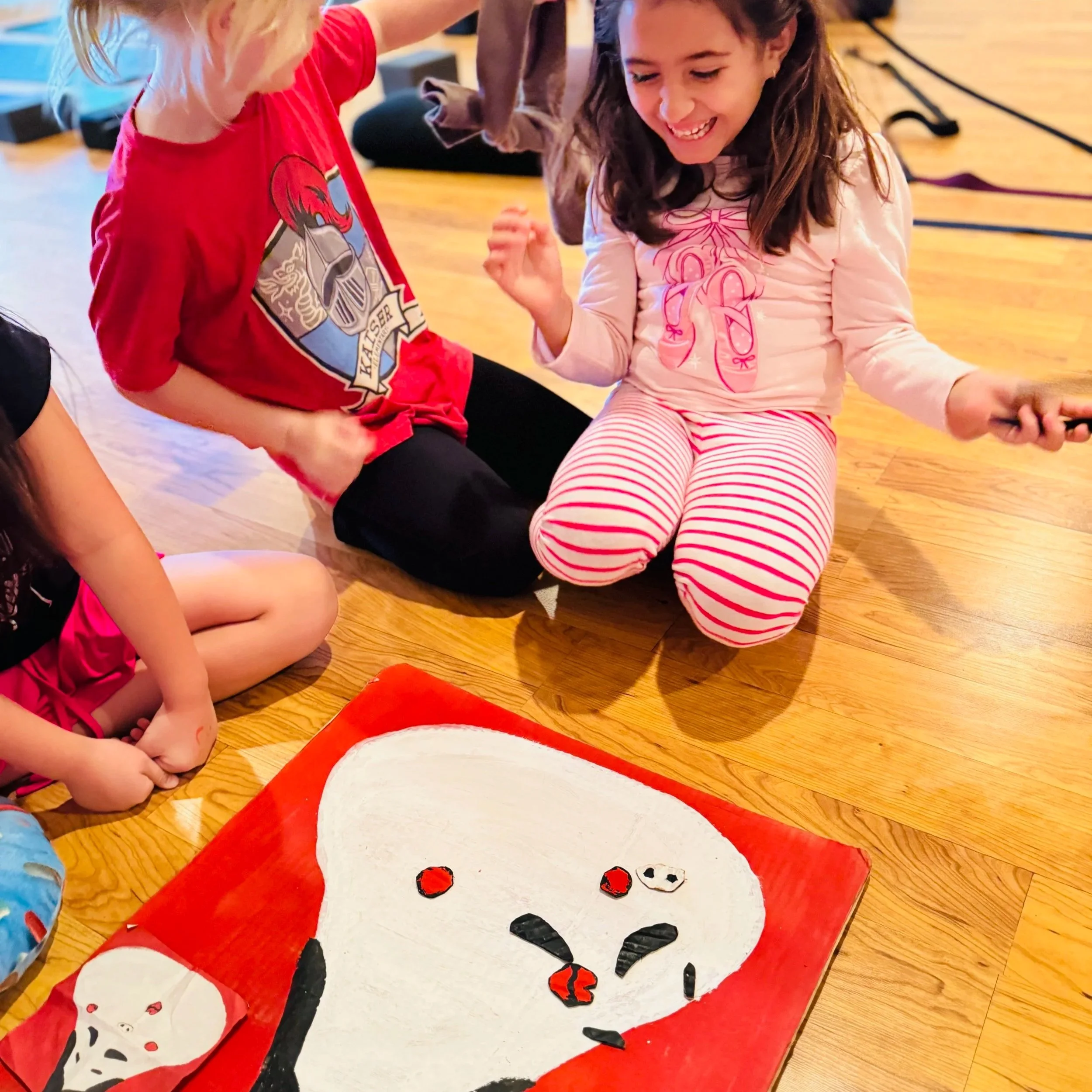 Children playing a game of pin the nose on the snowman on a wooden floor. The snowman is cartoon-style, painted on a large, flat surface, with a red background. One girl, with brown hair, is smiling and wearing a light pink shirt with a pink bow on i