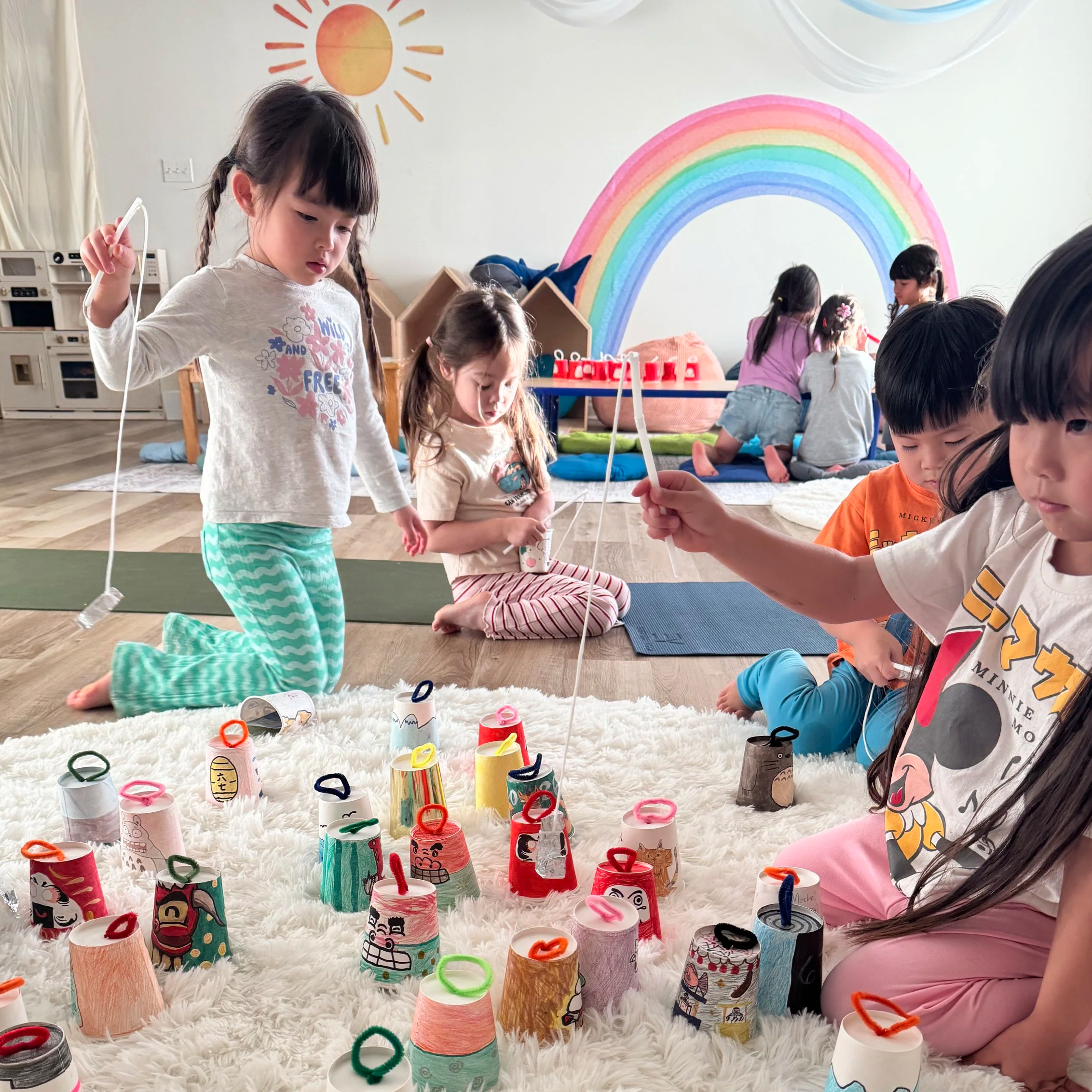 Children participating in a craft activity with decorated paper cups, sitting on a fluffy white rug in a room with a rainbow and sun painted on the wall.
