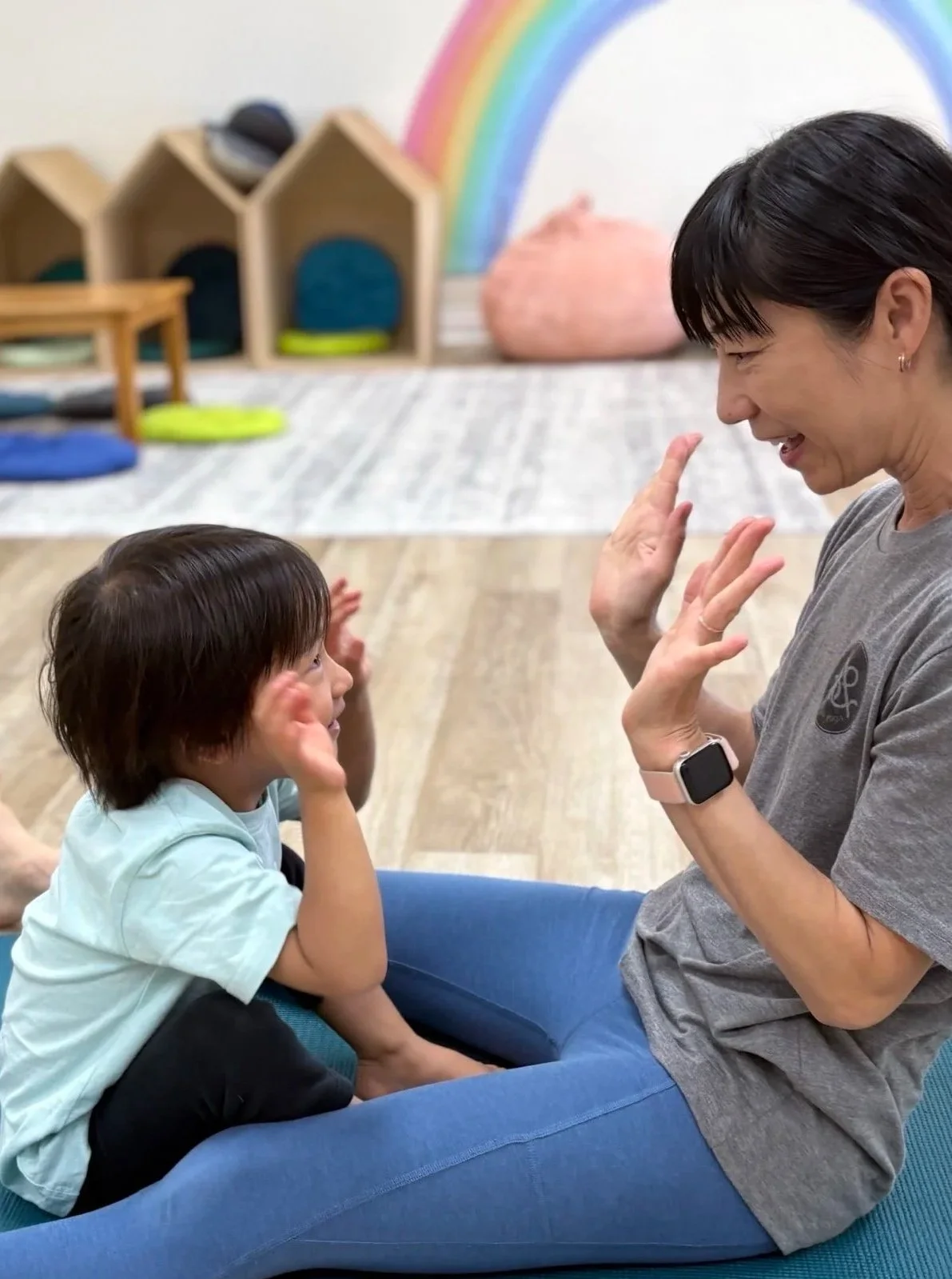 A woman and a child sitting on the floor facing each other, engaging in an interaction. The woman is smiling and has her hands raised in front of her face, while the child is looking up at her with a smile, touching his face with his hands. In the background, there is a colorful rainbow mural, small wooden houses with cushions inside, and a pink beanbag.
