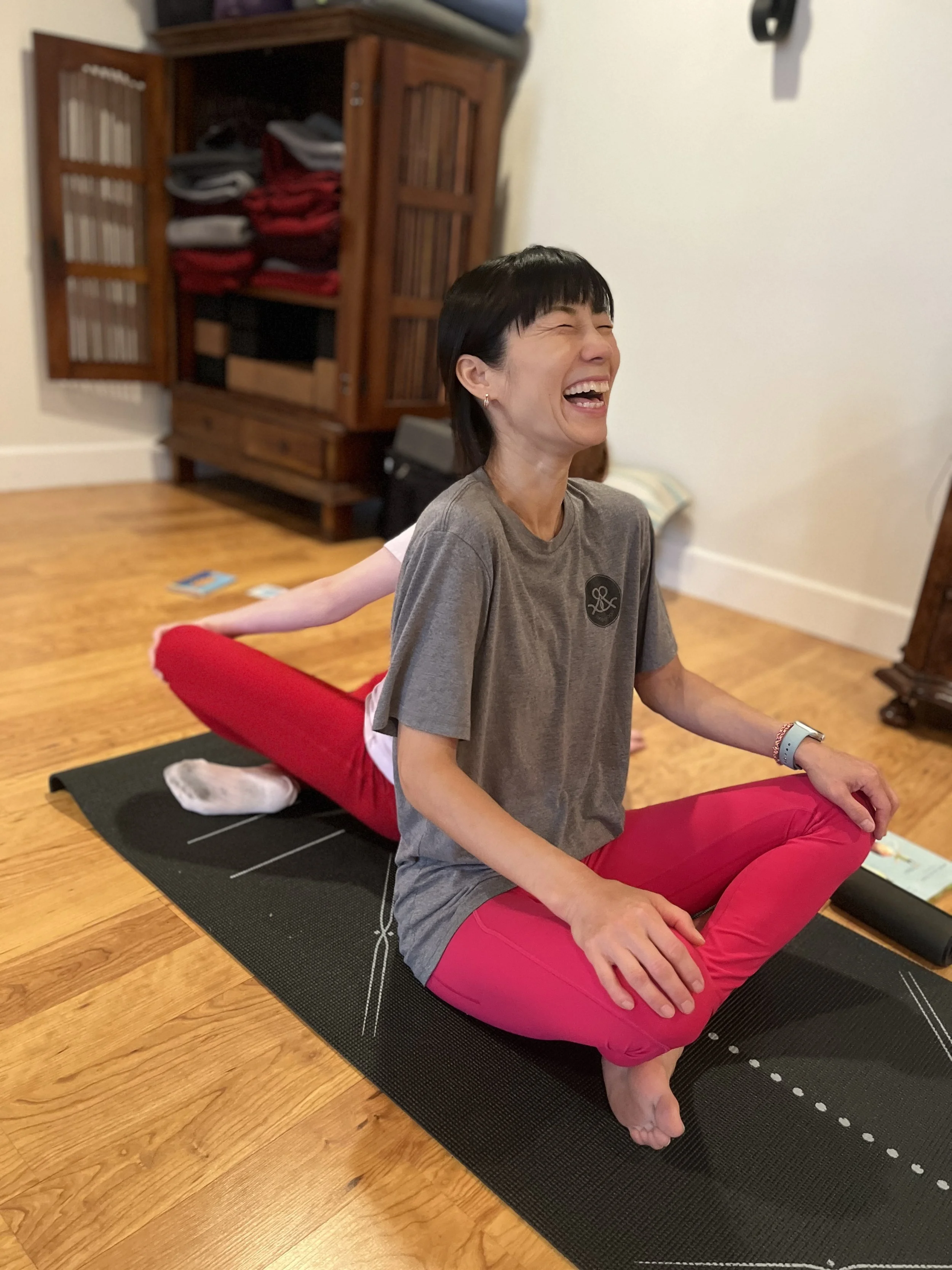A woman with short black hair and a big smile practicing yoga indoors on a black mat, sitting cross-legged on a red bolster with her hands resting on her knees. She is wearing a gray T-shirt and vibrant pink pants. In the background, there is a wooden cabinet with shelves filled with folded clothes and some books, along with wooden flooring.