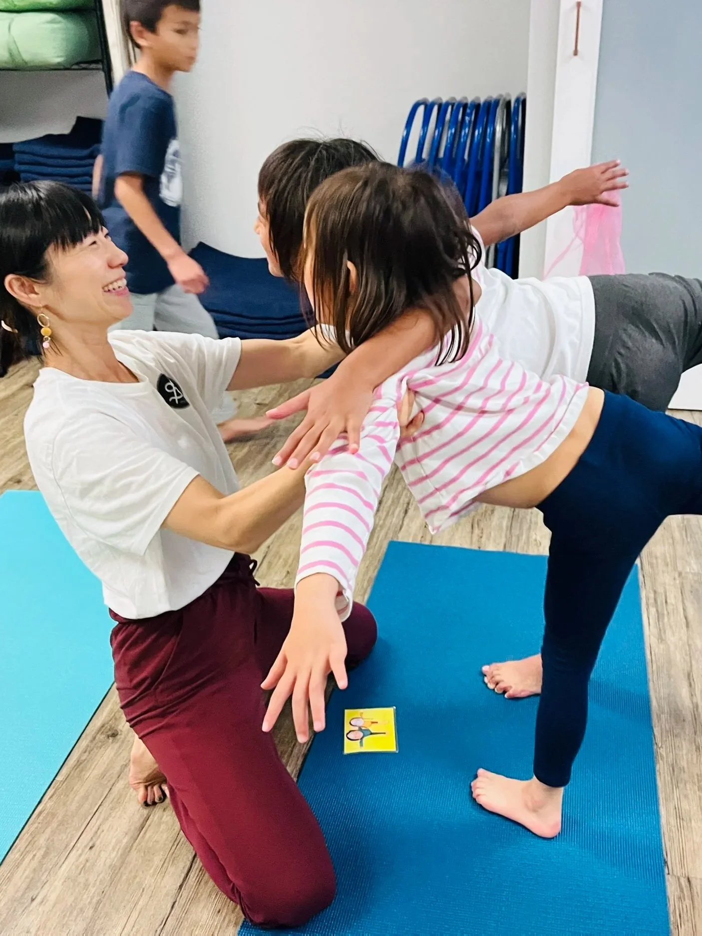 A woman and a young girl practicing yoga together indoors on yoga mats, with children and shelves in the background.