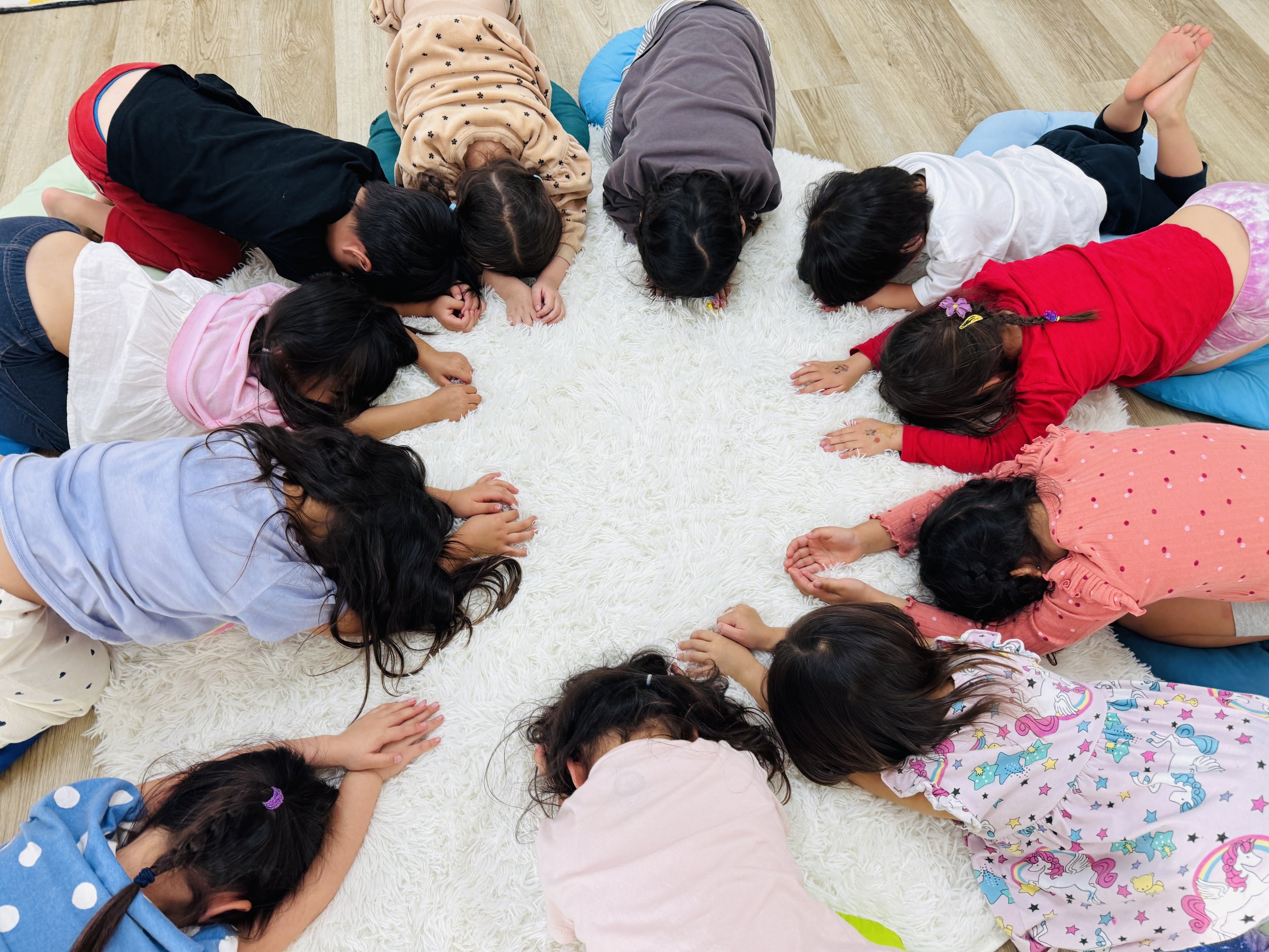 Children lying on a white shaggy rug on the floor with their heads close together in a circle, engaged in an activity.