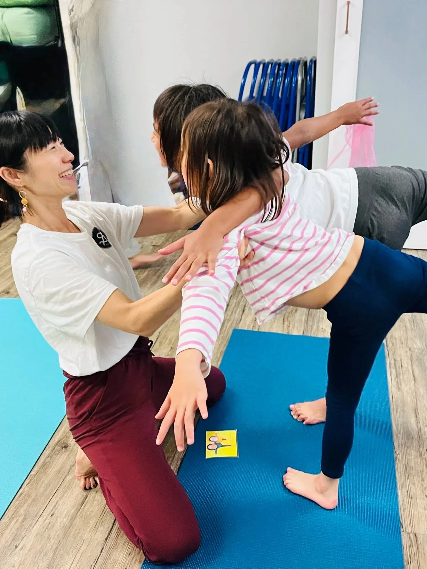 A woman helping two children with yoga poses on blue mats indoors. The woman is smiling and the children are balancing on a yoga mat with their arms extended, practicing yoga together.