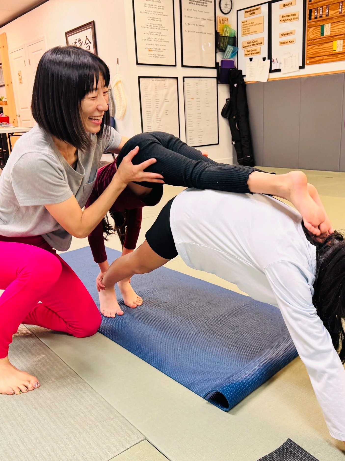 A woman is assisting a person with a forward bend yoga pose during a class. The woman is smiling and supporting the person at hips and knees, with a yoga mat on the floor. The background shows a whiteboard with schedules and notices on the wall.
