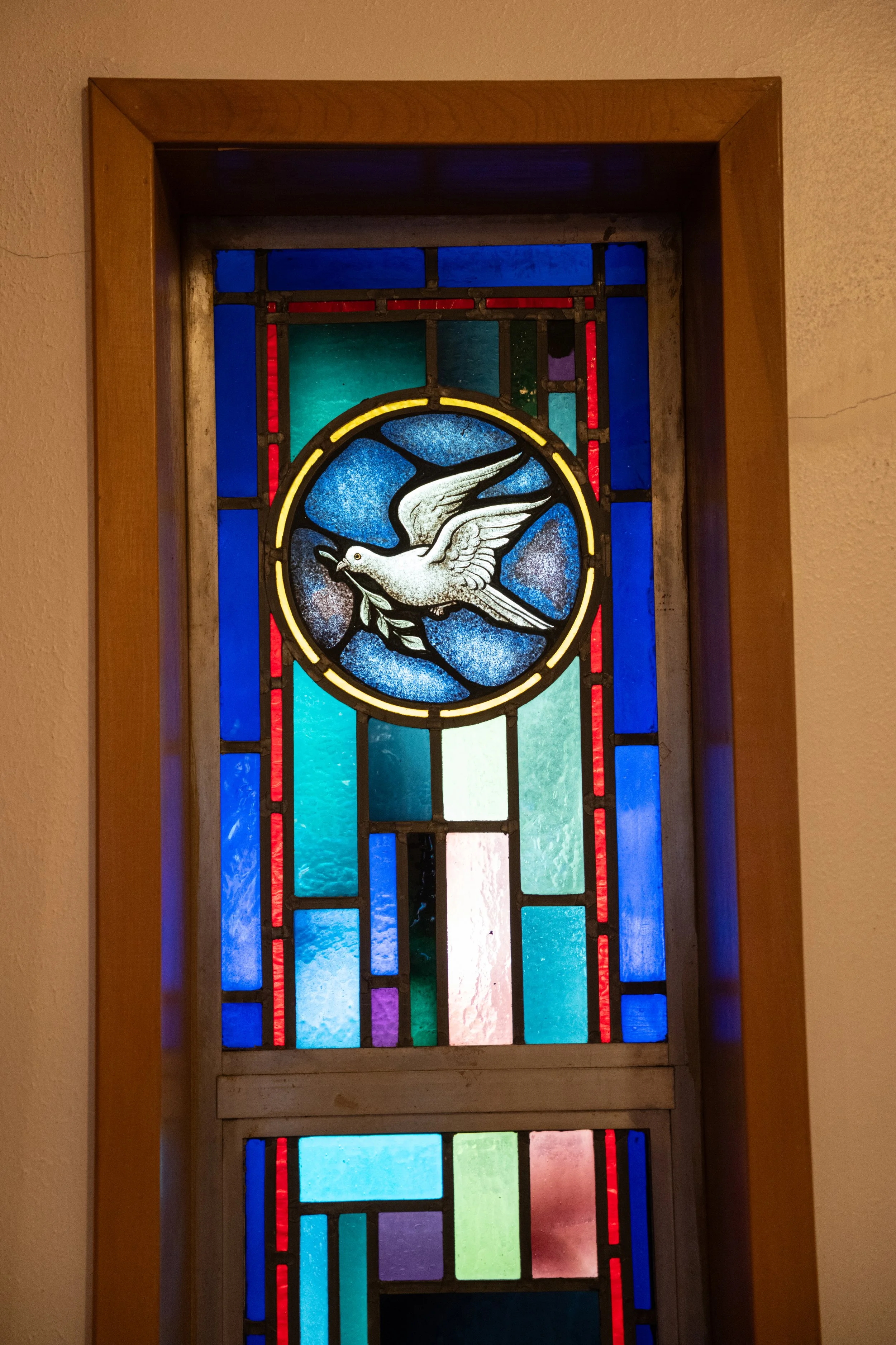 Colorful stained glass window with a white dove carrying an olive branch at the center, framed by a wooden border.