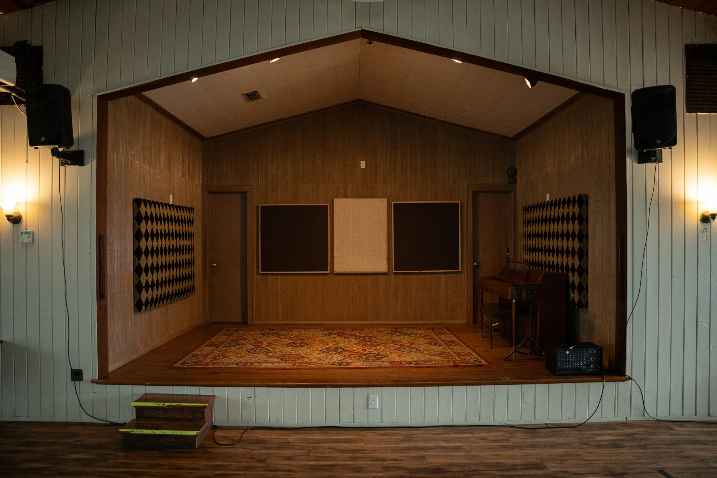 An empty small stage with a wooden floor and a rug, featuring a decorative black and gold acoustic panel on each side, with black walls and a beige ceiling with spotlights, a piano, and audio equipment.
