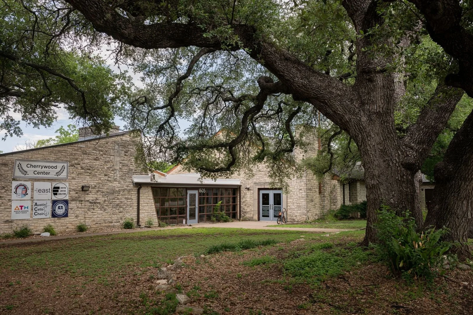 A stone building labeled Cherrywood Center with a large tree in front of it, a small bicycle parked near the entrance, and a grassy area with a few small plants.