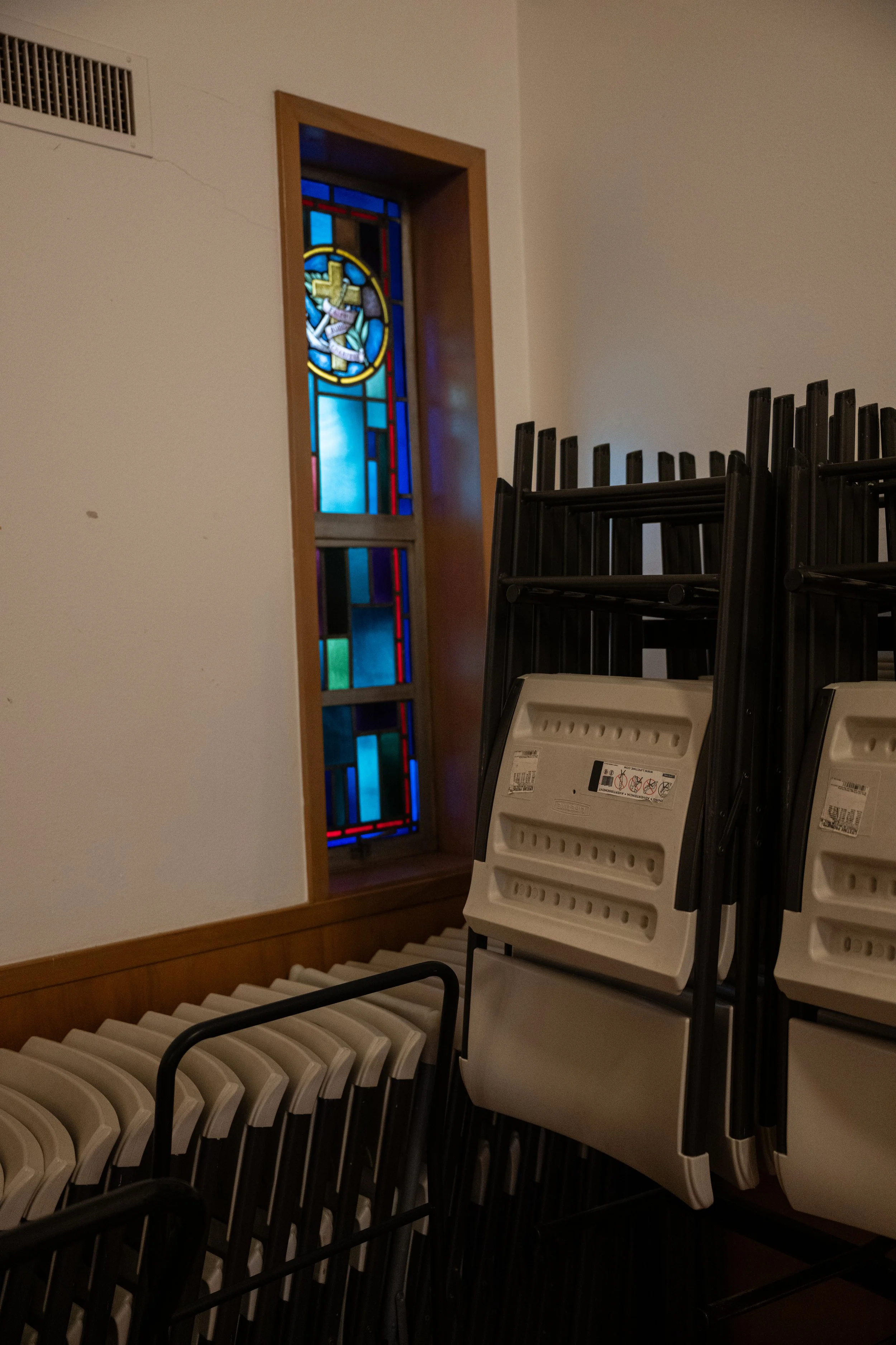 Church interior with stacked white plastic chairs, a stained glass window depicting a religious figure, and a wall vent.