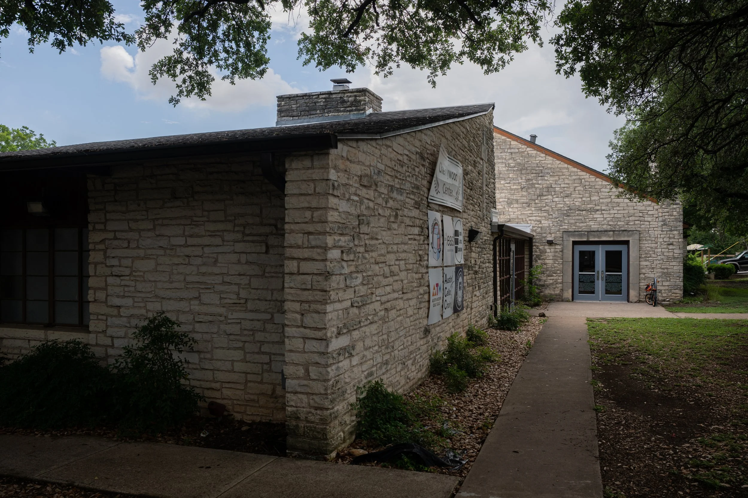 A brick building with signs on the wall, a paved sidewalk, and trees overhead. The sky is partly cloudy.