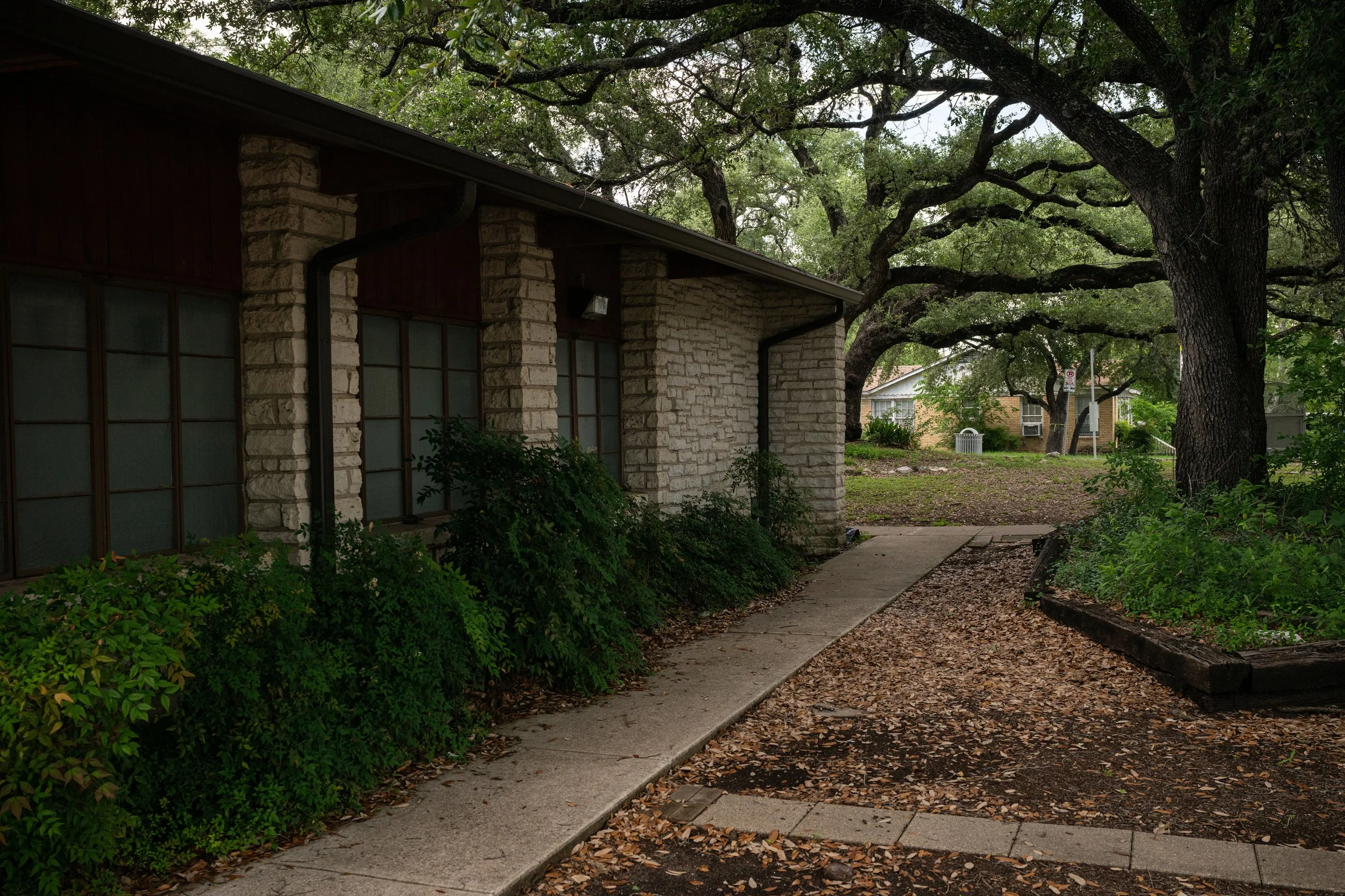 Sidewalk running past a building with stone columns, surrounded by trees with overhanging branches in a residential neighborhood.