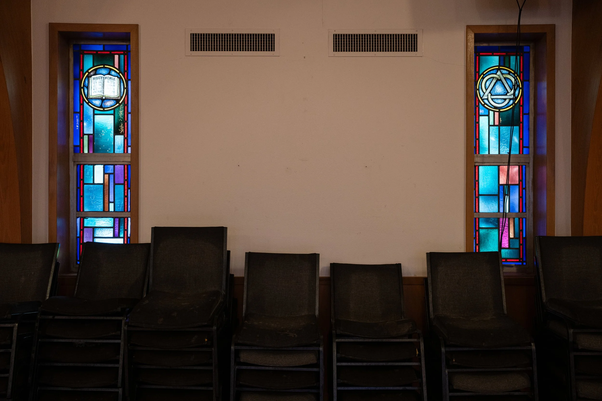 Interior view of a church with two stained glass windows on a plain wall, and empty black chairs lined up in rows in the foreground.