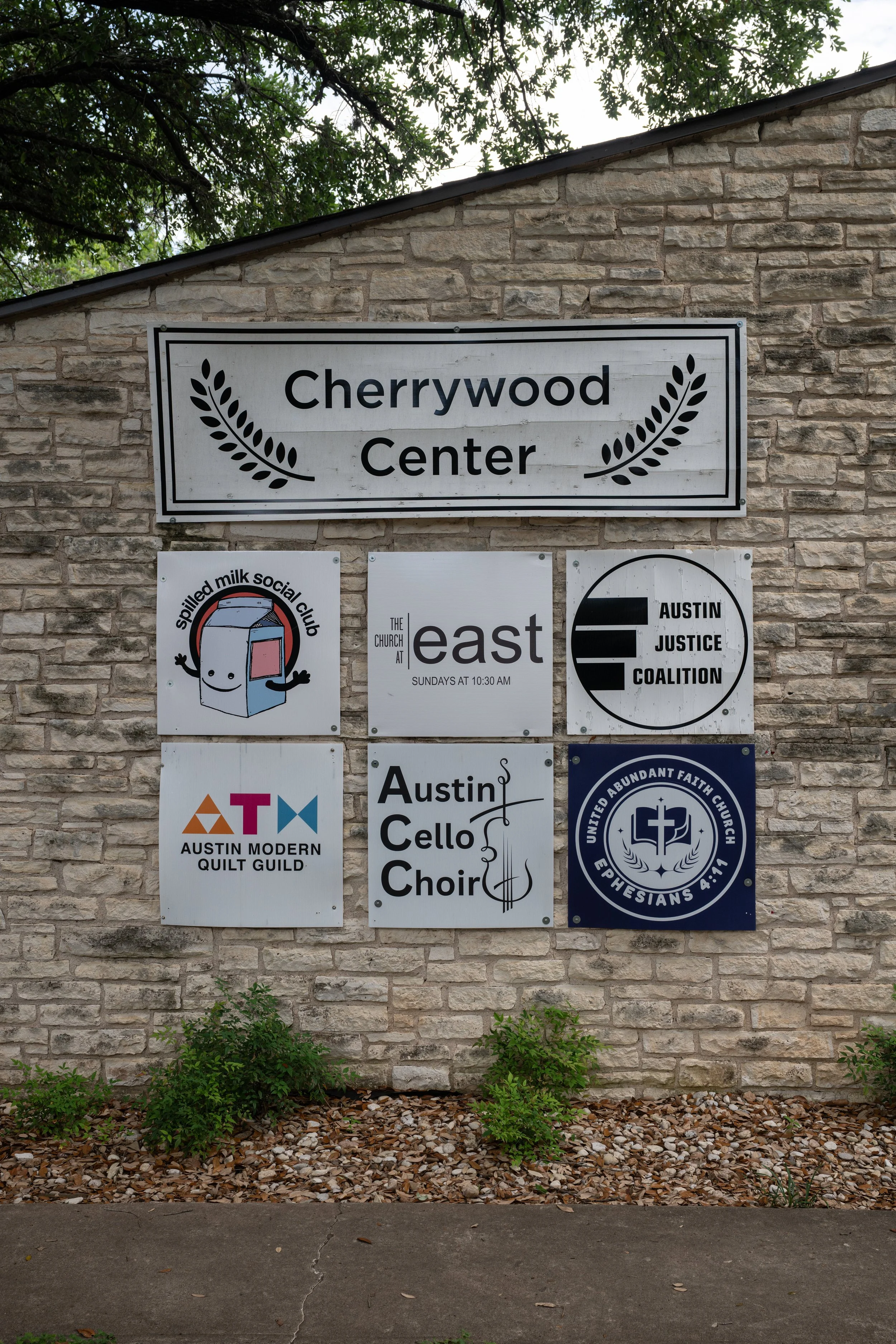 A brick wall with a large sign reading 'Cherrywood Center' and smaller signs below for various community organizations, with some greenery and a sidewalk in the foreground.