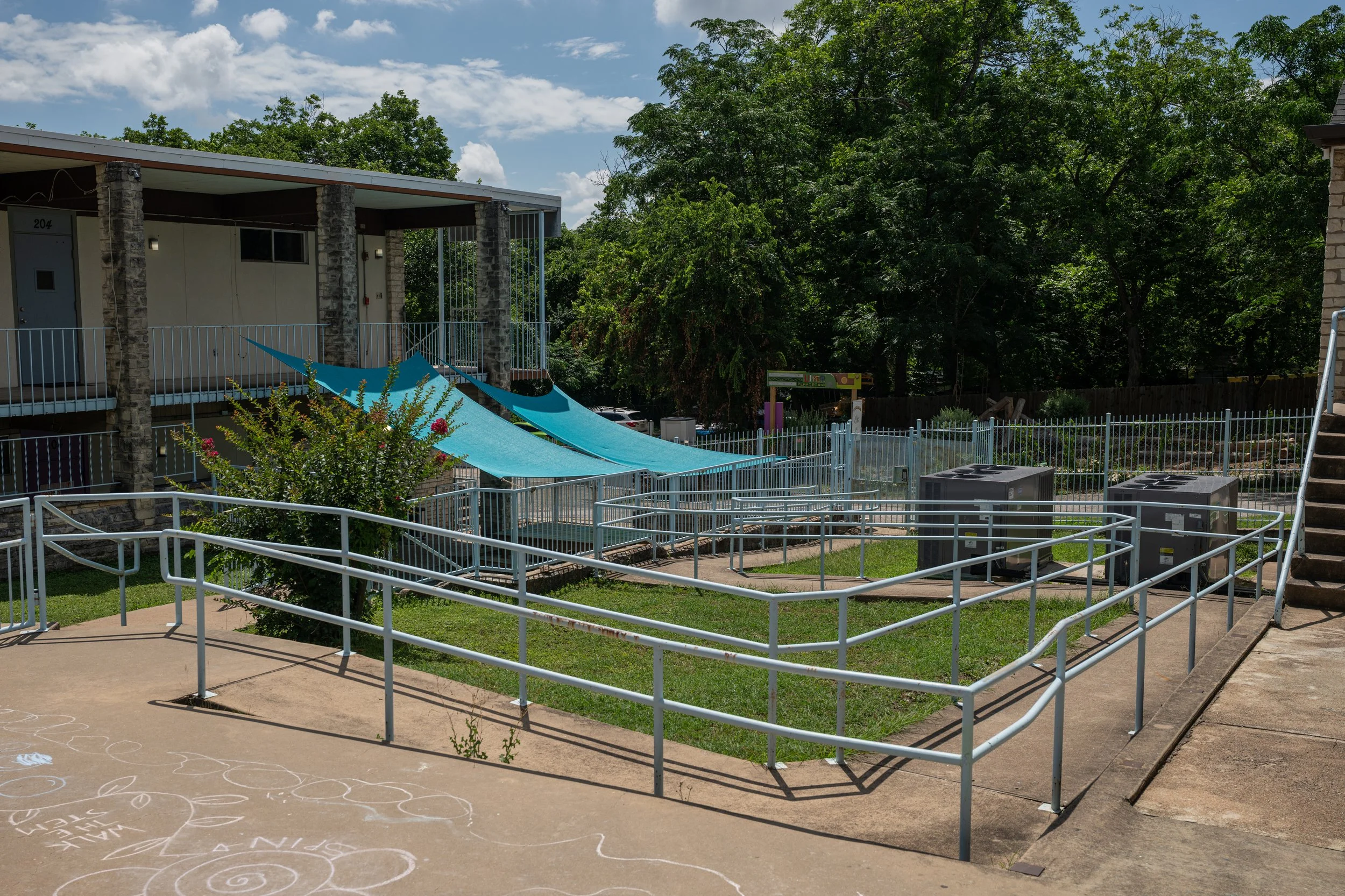 An outdoor scene with apartment buildings, lush green trees, and a clear blue sky with white clouds. There are blue shade sails over a playground area with metal fencing, and chalk writings on a concrete pathway.