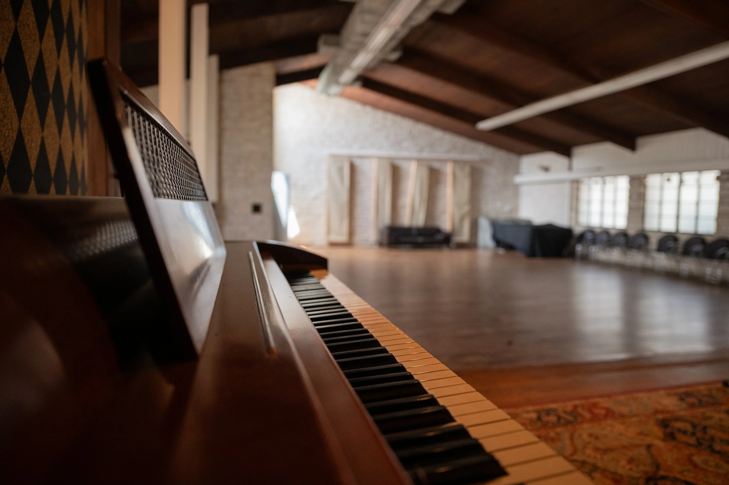 A close-up of a piano keyboard in a spacious room with wooden floors, exposed wooden ceiling beams, large windows, and a seating area with black couches in the background.