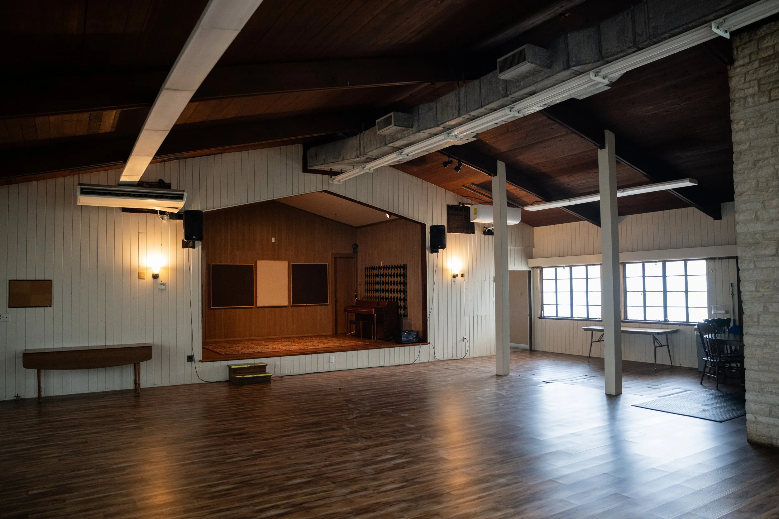 Empty interior of a room with wooden floors and a stage, featuring a few chairs, a black speaker, and wall-mounted lights.