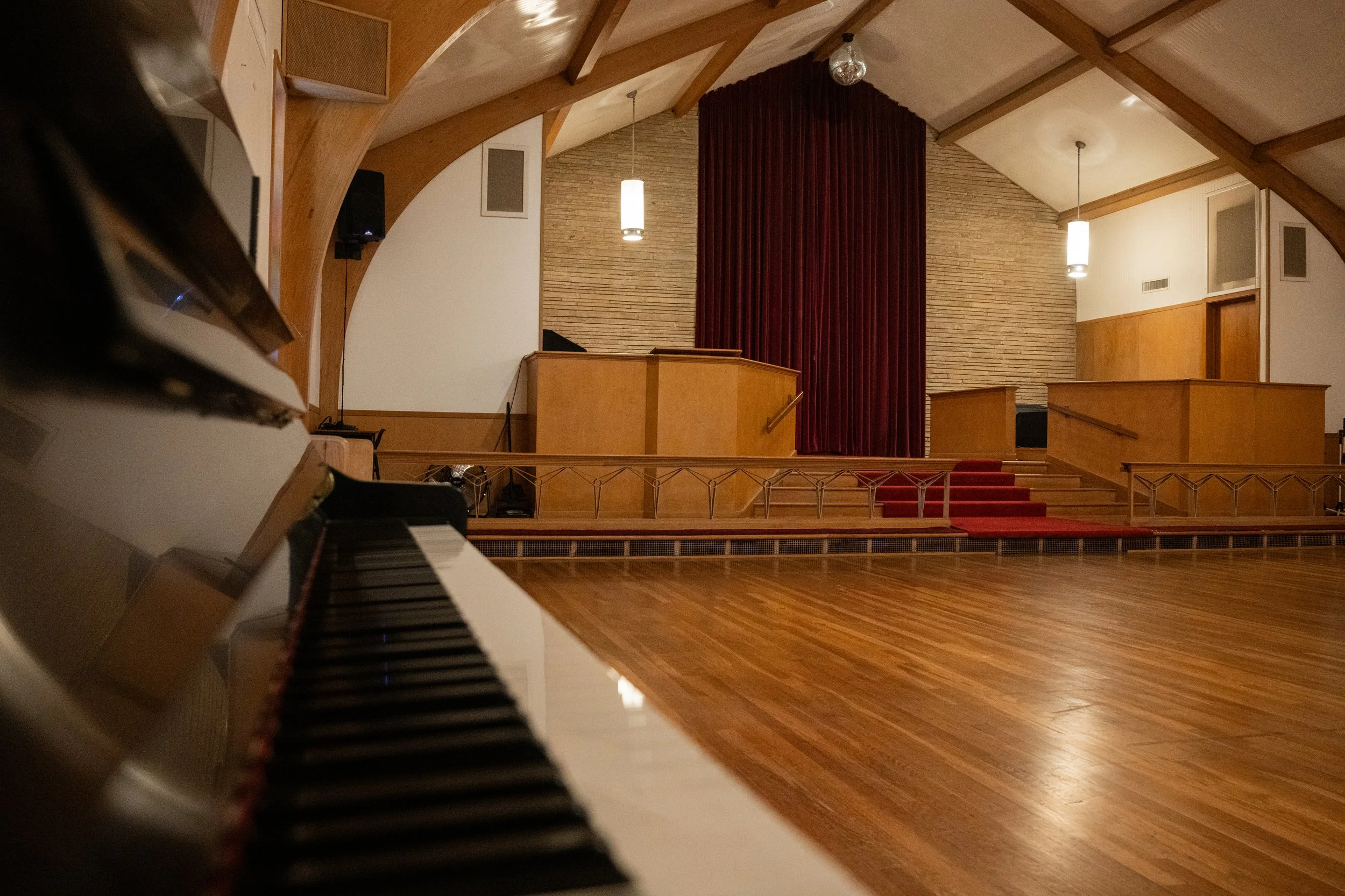 Interior of a music performance space with wooden accents, stage, red curtain, pendant lights, and a grand piano in the foreground.