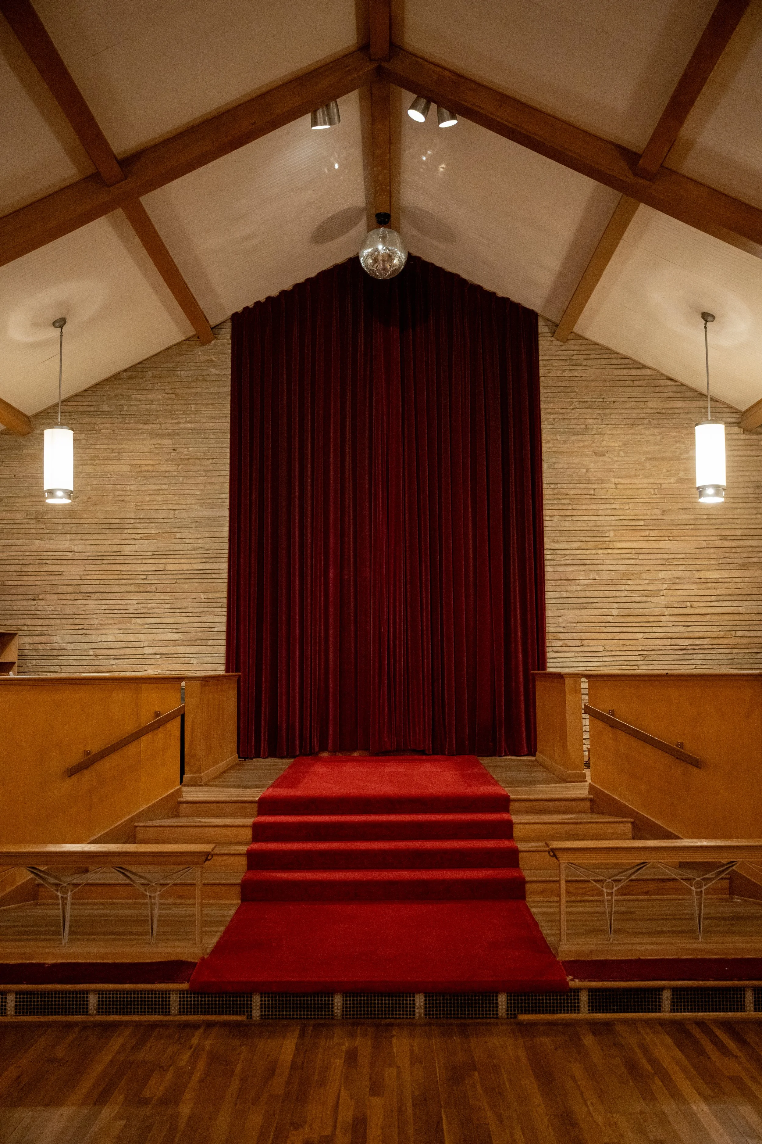 An interior view of a church or auditorium stage with a red carpeted altar, surrounded by wooden railings, and large red curtain behind it. The ceiling features wooden beams, a disco ball, and hanging pendant lights.
