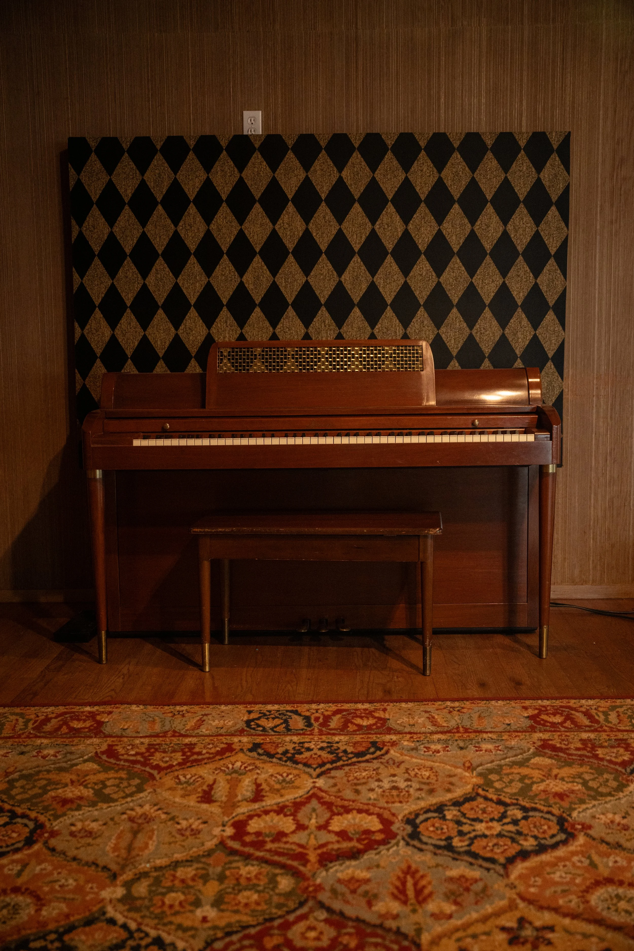A vintage upright wooden piano in a dimly lit room, with a wooden wall background and a decorative area rug on the floor.