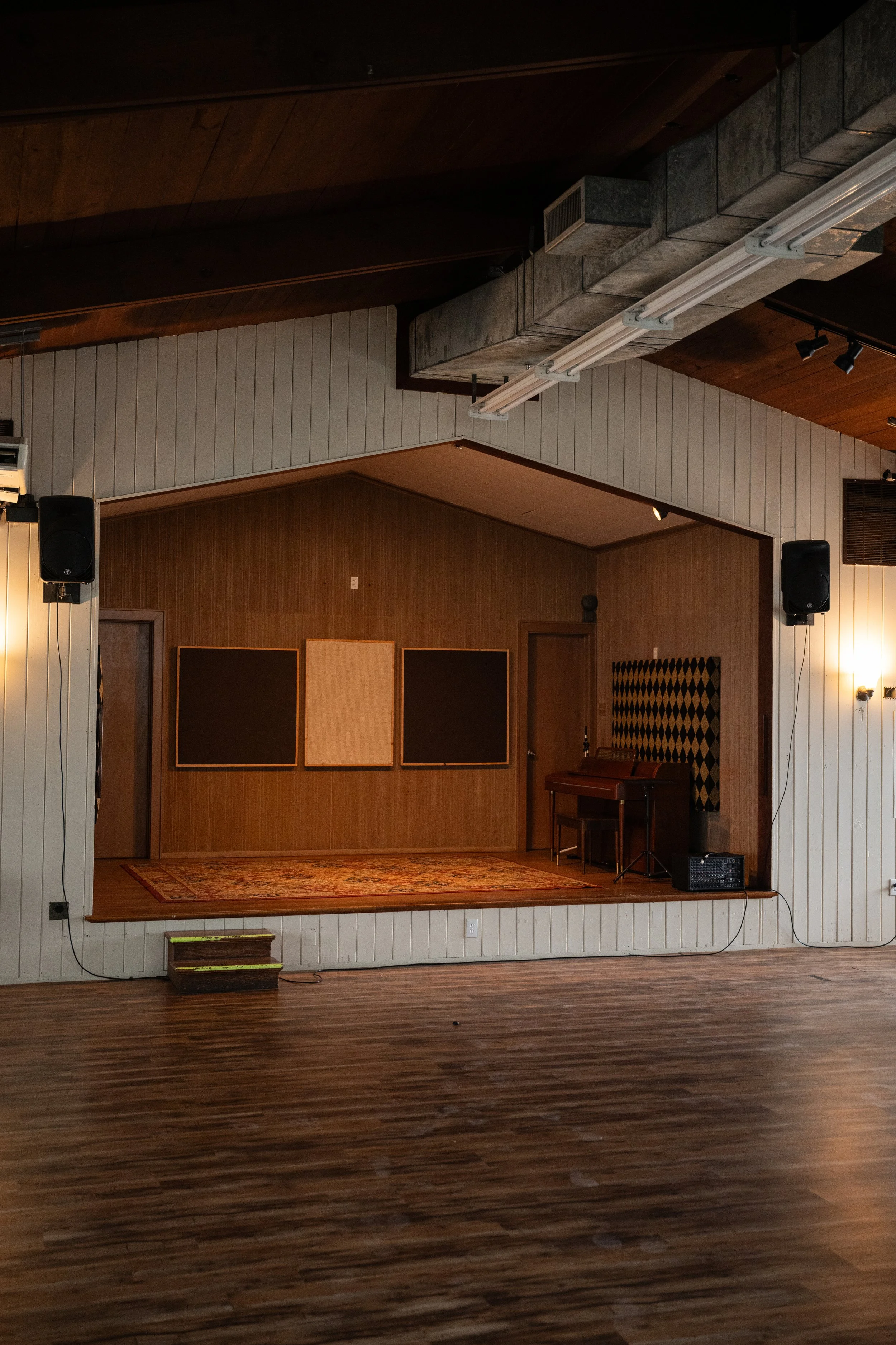 Empty stage in a small performance hall with wooden walls and flooring, featuring a rug, a piano, folder on a stand, speaker, and sound equipment.