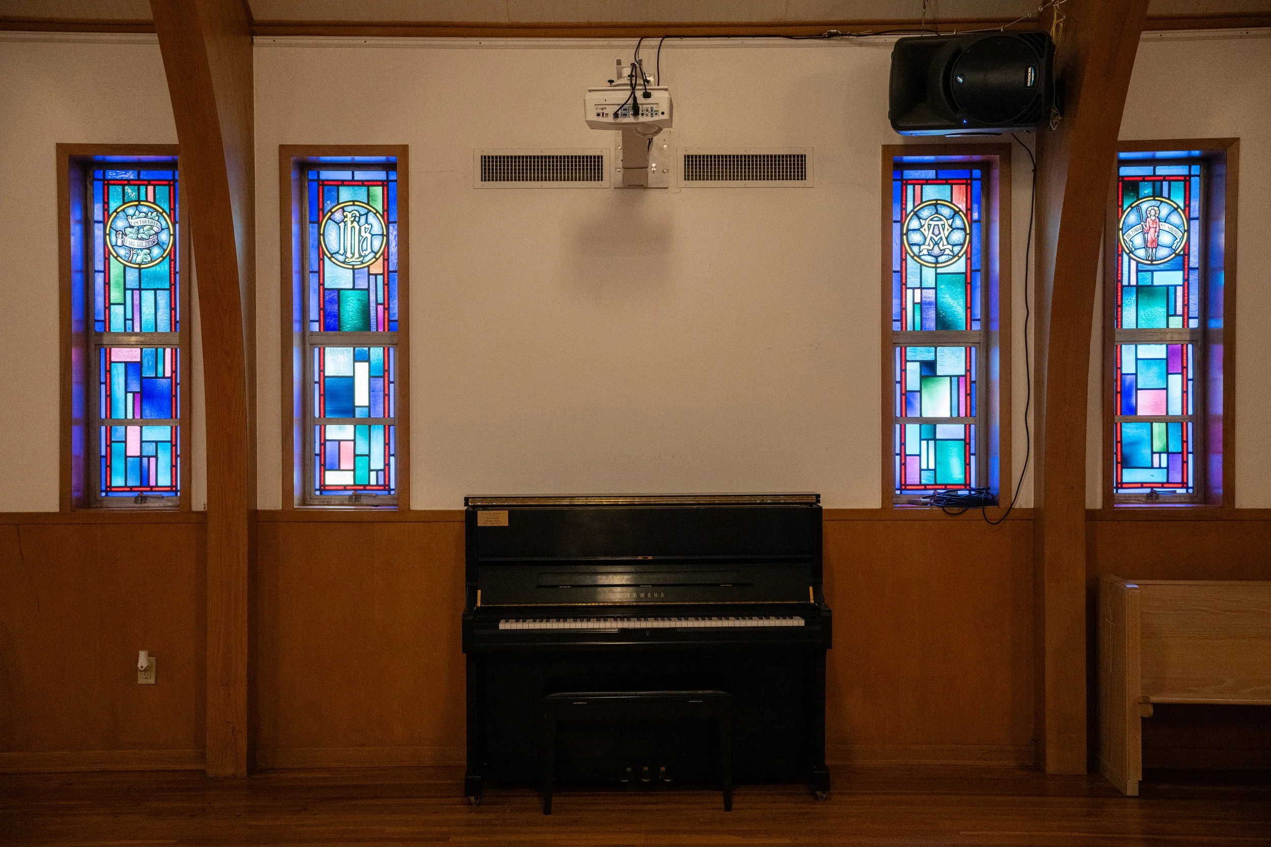 Interior of a room with four stained glass windows, a black upright piano in the center, a wooden beam to the left, and a large black speaker on the wall to the right.