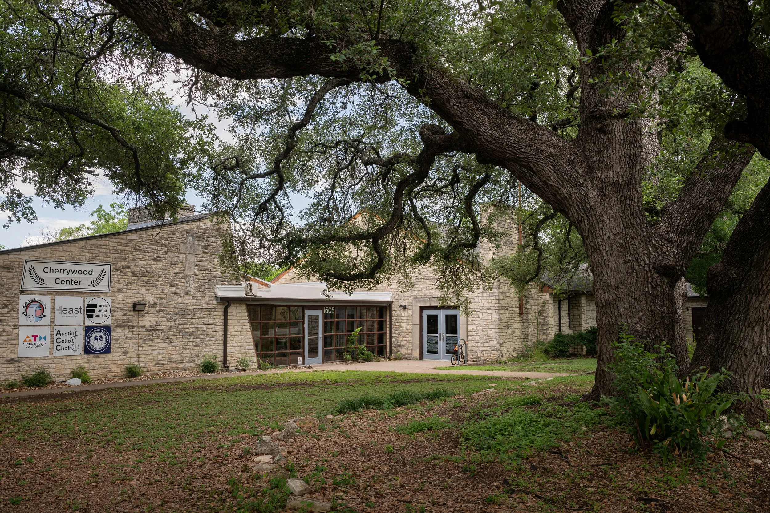 A large tree with sprawling branches and textured bark stands in front of a stone building with a sign indicating it is the Cherrywood Center. The building has multiple logos and signs on its exterior, and a bicycle is parked near the entrance.