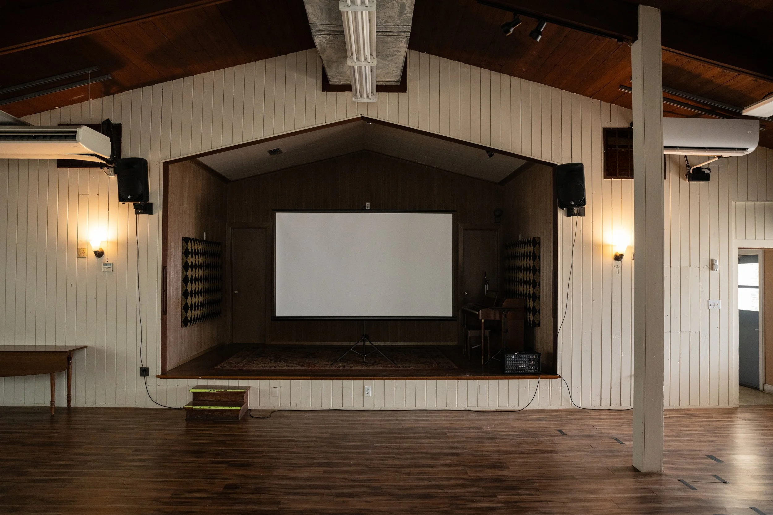 Meeting room with a large projection screen on the wall, surrounded by wooden panels, with audio speakers, air conditioning units, and lighting fixtures.