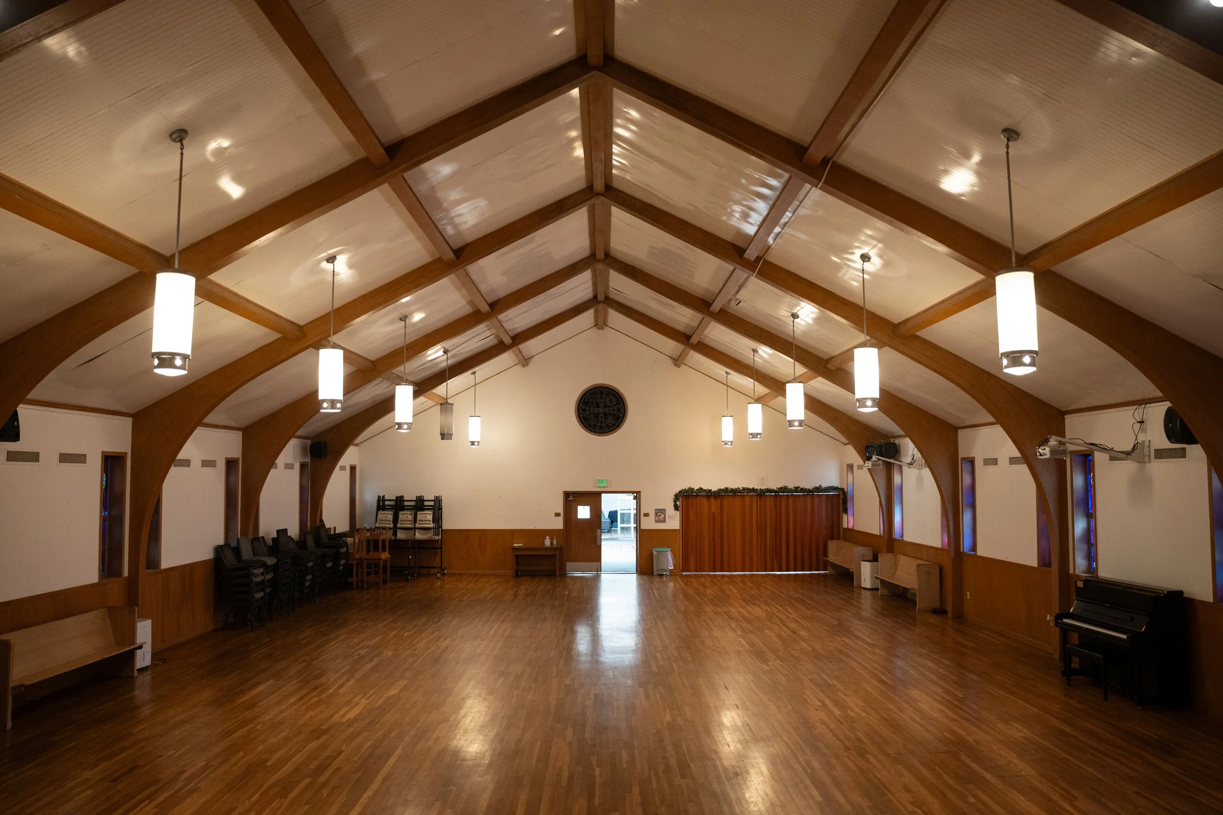 Empty church hall with wooden beams on the ceiling, hanging pendant lights, and a hardwood floor. Chairs are stacked against the wall, and a piano is visible on the right side of the room.