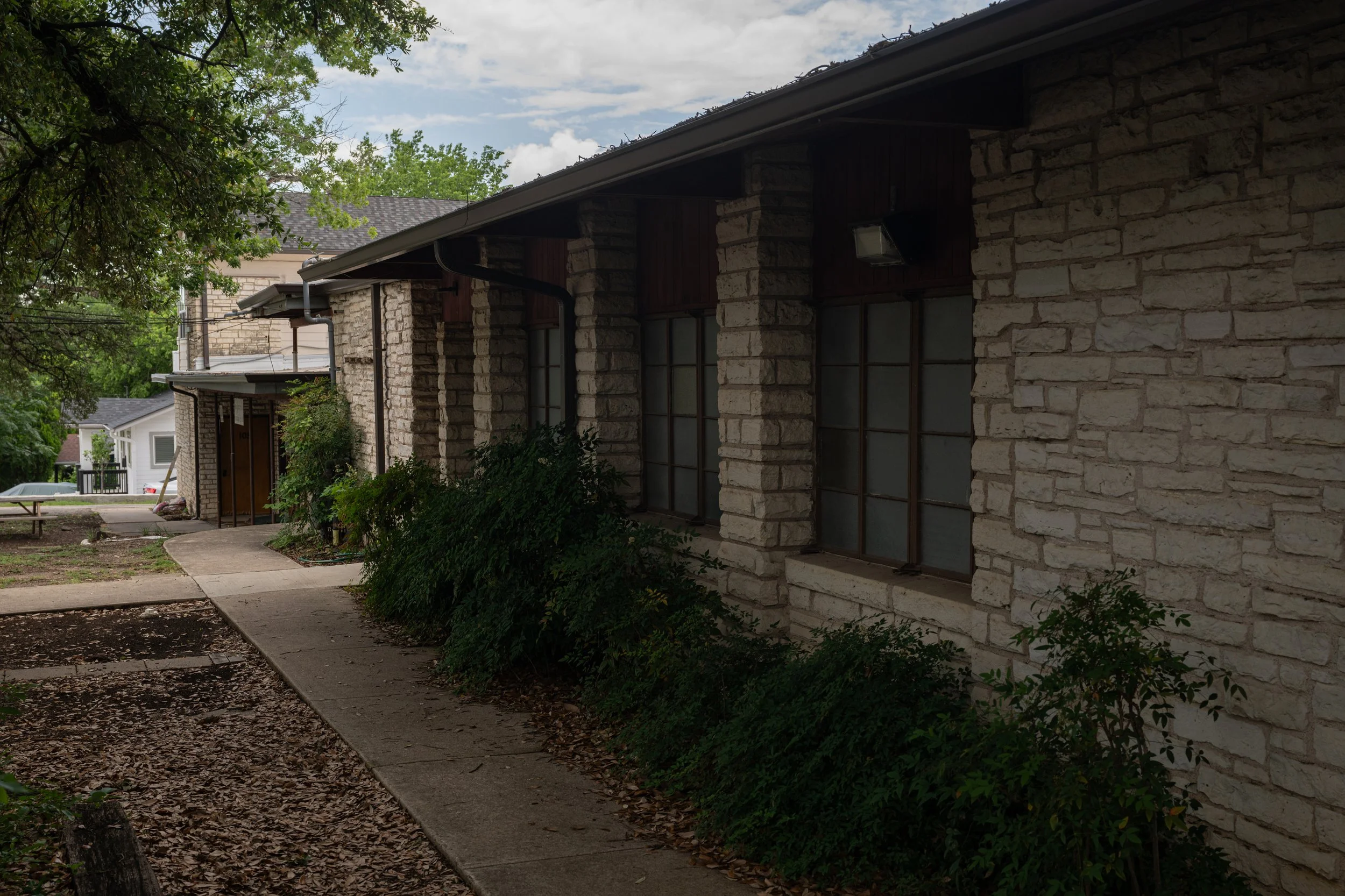 A sidewalk runs alongside a stone house with multiple windows, green plants, and a tree overhead, under a cloudy sky.