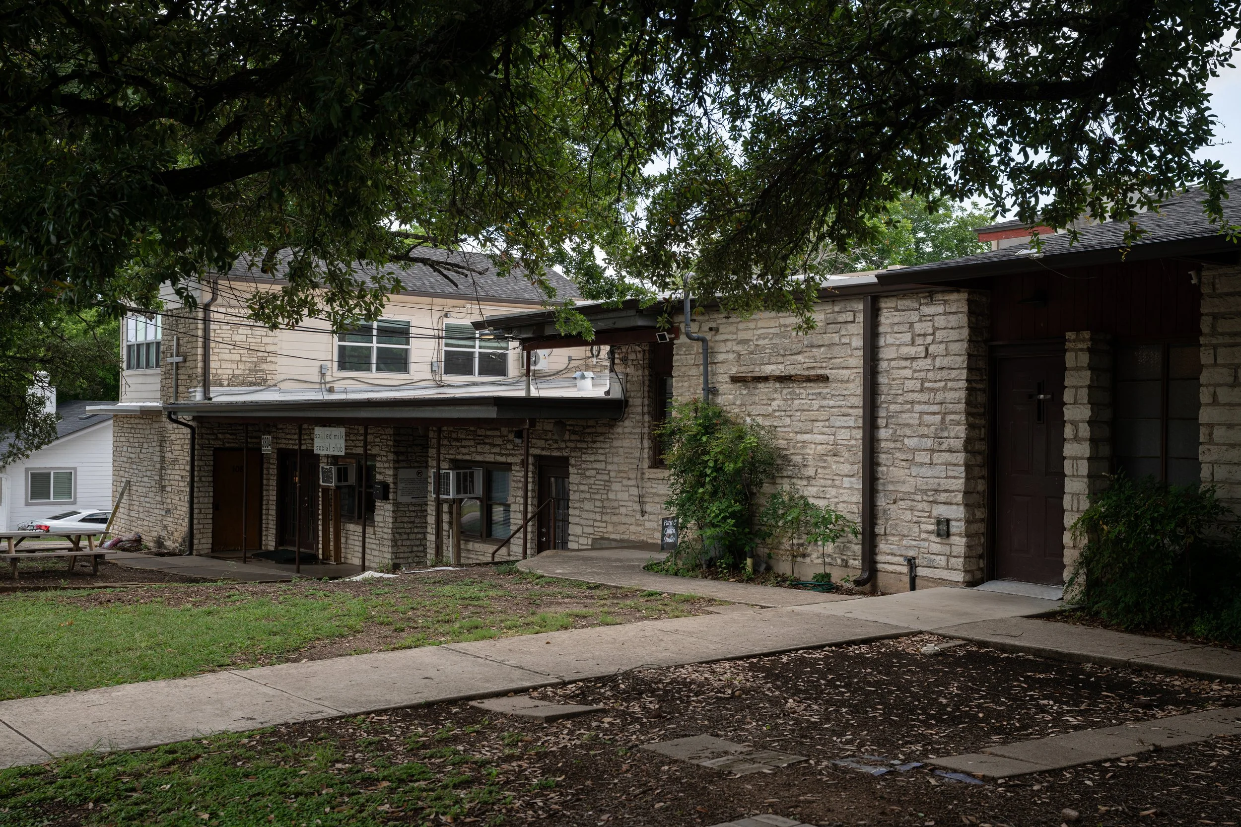 A residential building made of stone and brick with large windows, partially obscured by tree branches and foliage in a yard with a sidewalk and patches of grass.