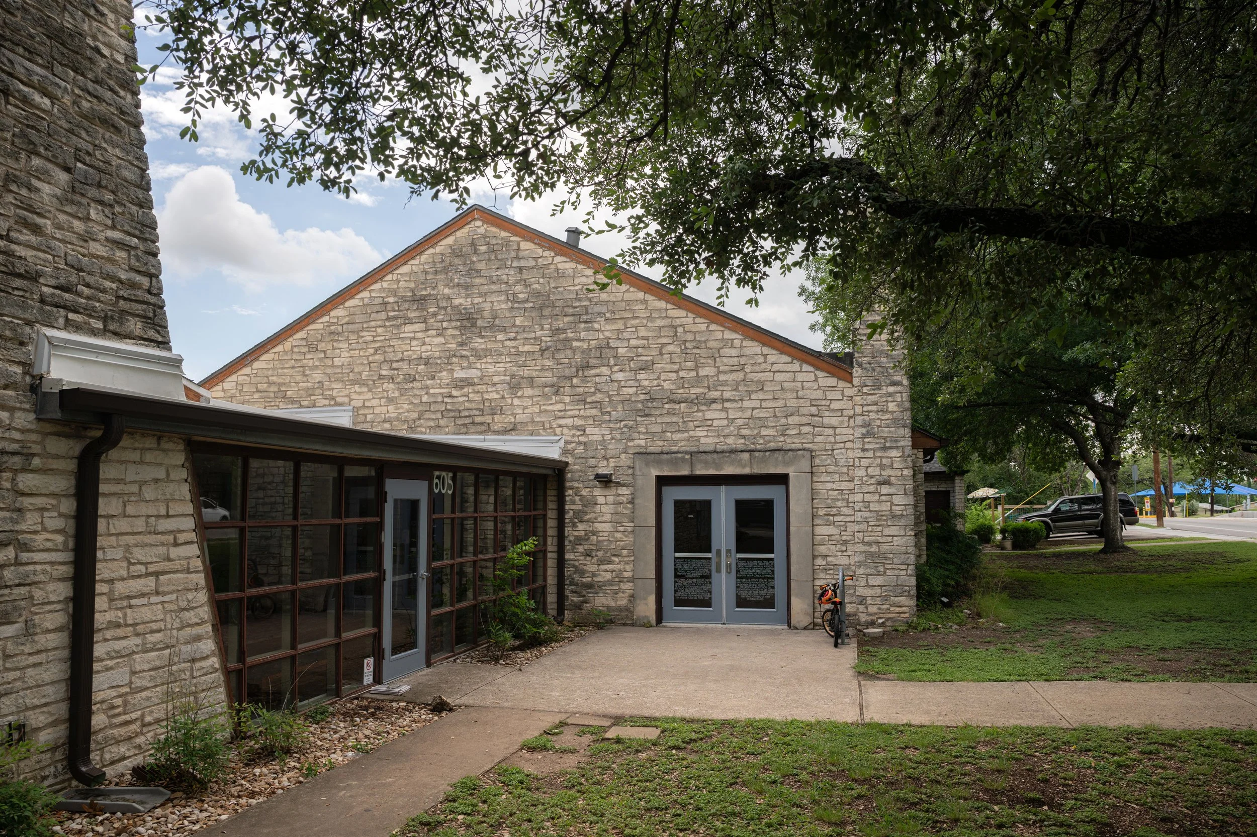 Front of a beige brick building with glass doors, a large window, a small tree, and a concrete walkway. Trees and cars are visible in the background under a partly cloudy sky.