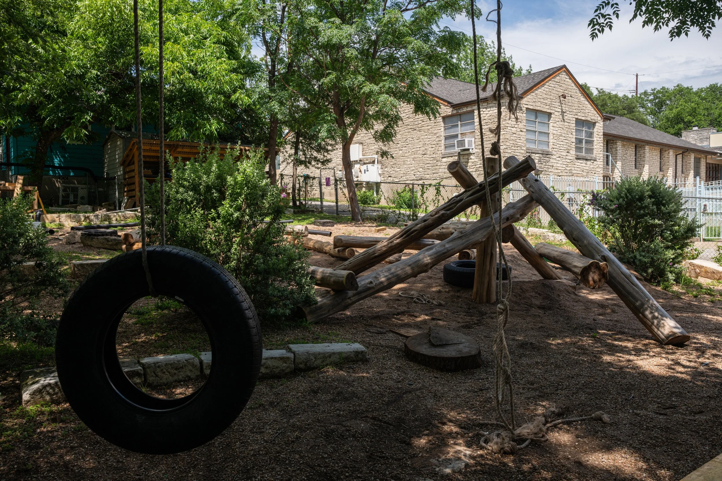 Backyard with trees, a wooden swing set, tire swings, and a house with stone walls and multiple windows.