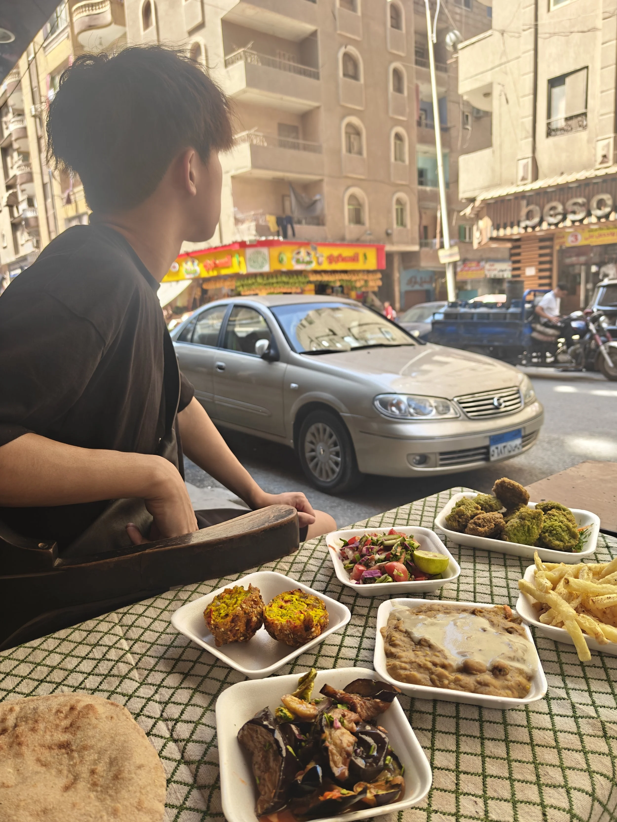 A table with various dishes of food, including vegetable fritters, roasted eggplant with toppings, a vegetable salad, a creamy dish, fried potato strips, and bread, set outdoors with a young man sitting nearby on a busy city street.