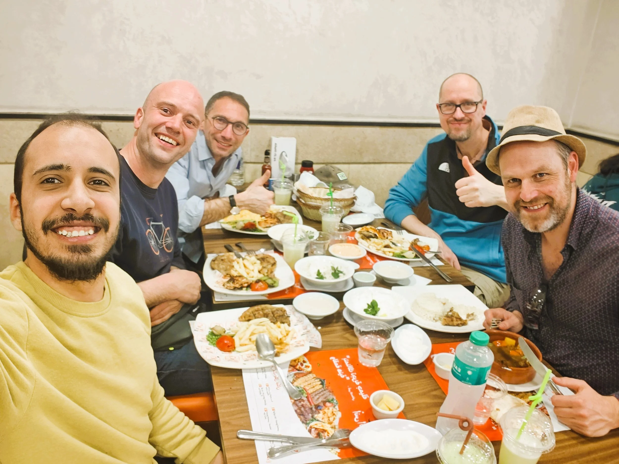 A group of five men sitting at a dinner table in a restaurant, smiling and taking a selfie. The table is filled with various dishes, drinks, and condiments.