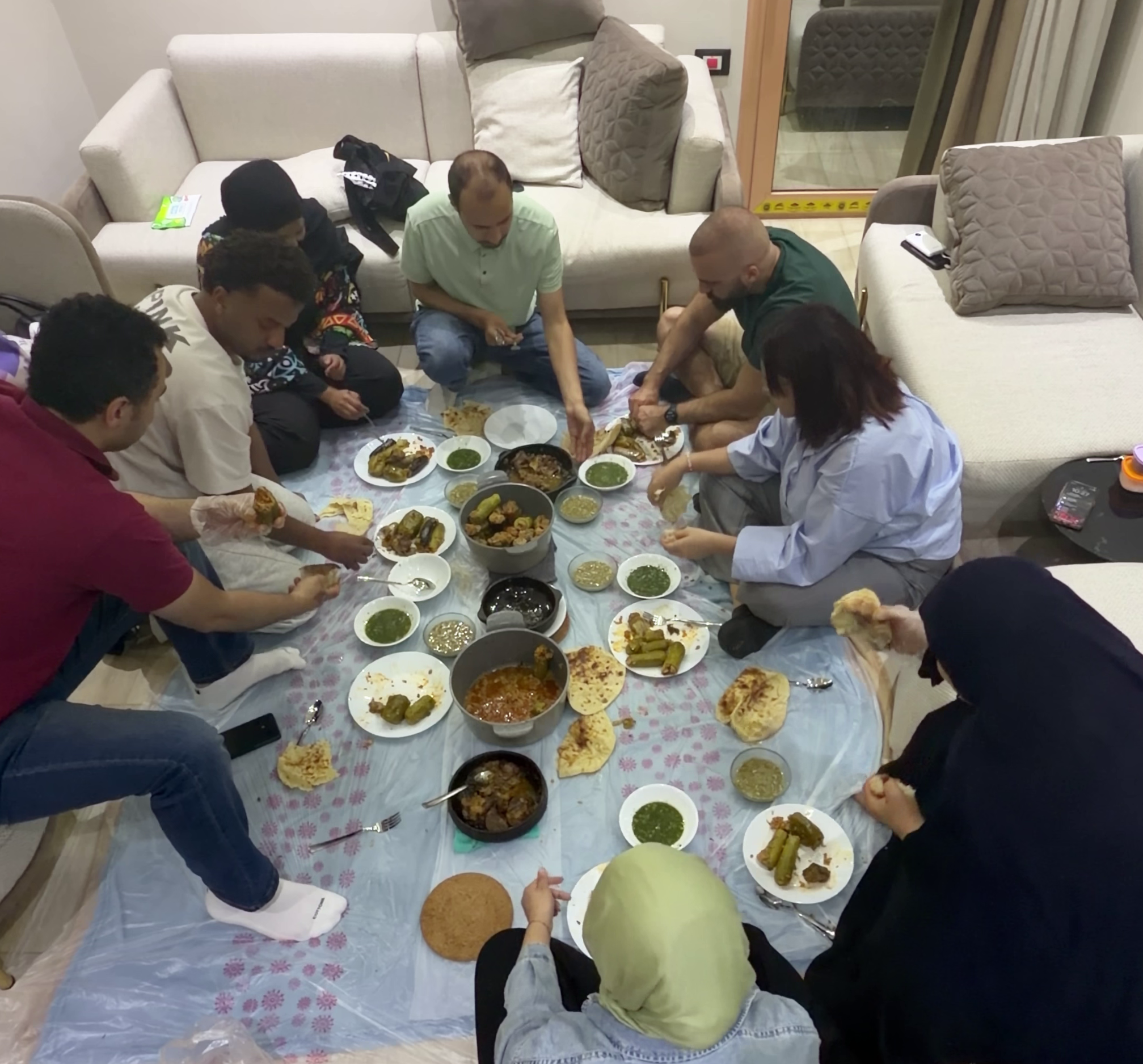 A group of diverse people gathered around a low table on a cloth, eating various dishes of Mexican cuisine such as tacos, enchiladas, and soups, in a cozy living room with sofas and pillows.