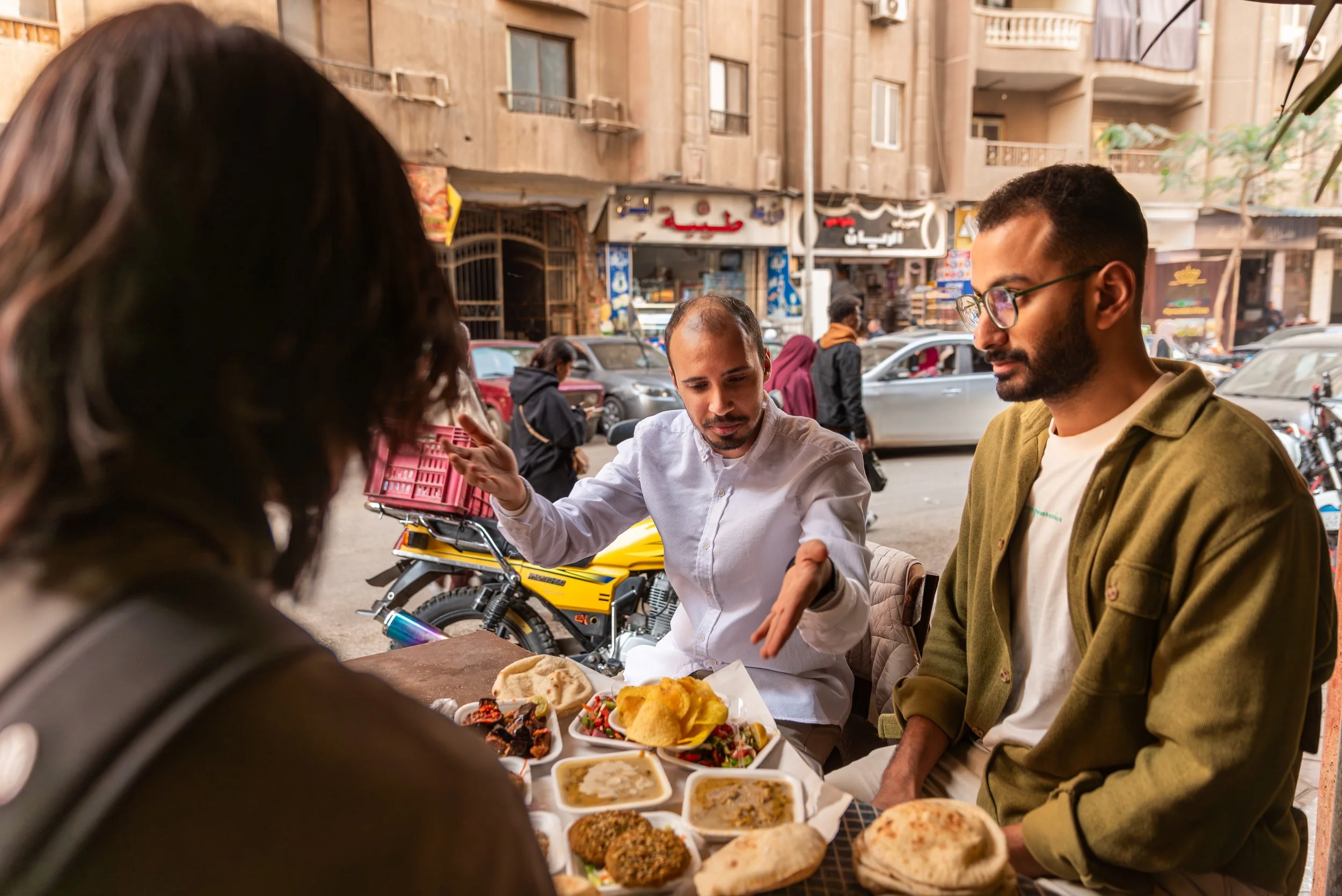 People at an outdoor market or food stall. Two men are visible, one with glasses and a beard, the other with a shaved head, both engaged in food tasting or ordering. Plates of food, including bread, vegetables, and other dishes, are on the table. The background shows a busy street with cars and shops.