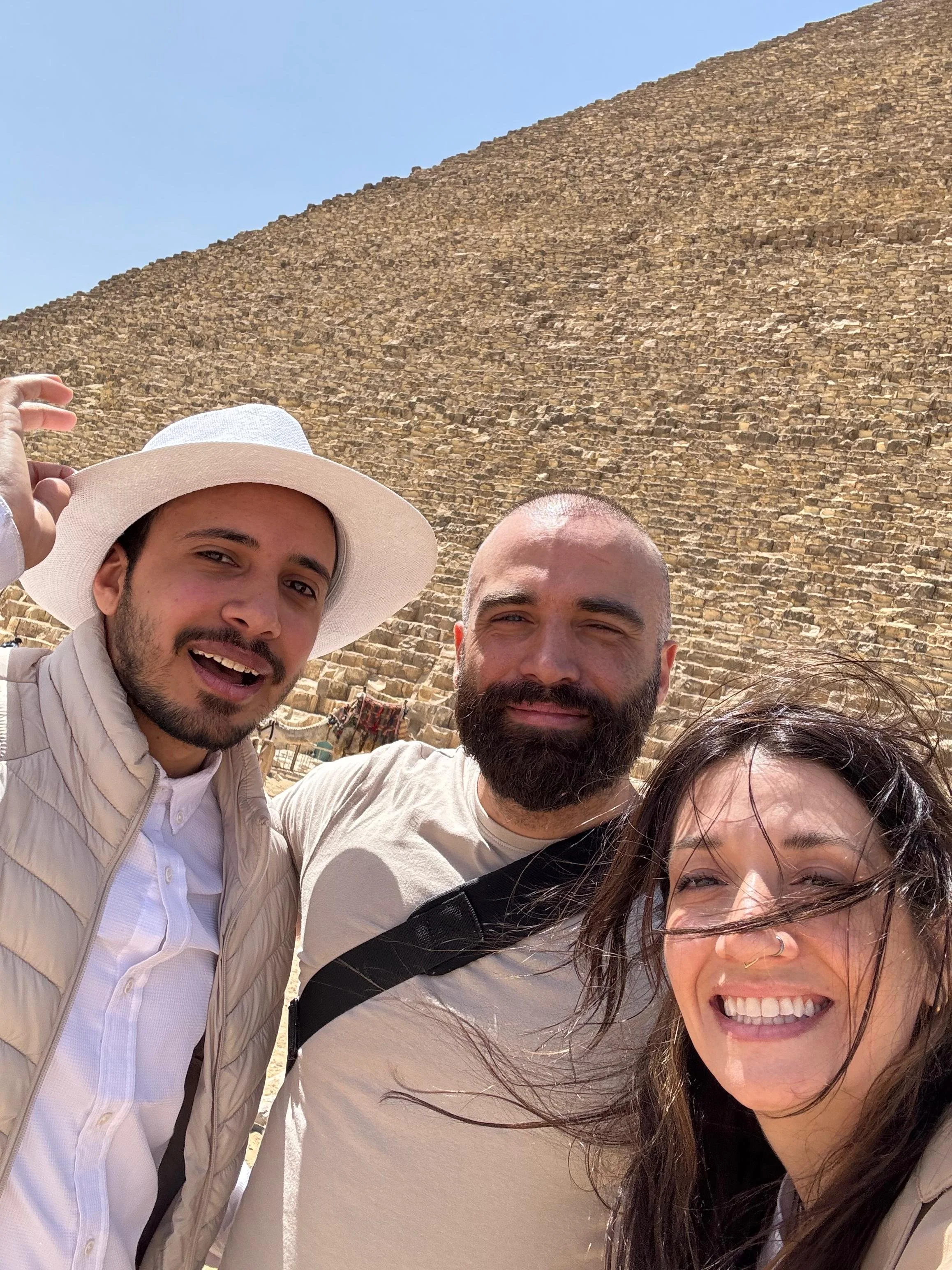 Two men and one woman taking a selfie in front of the Great Pyramid of Giza under a bright blue sky. The woman has long hair and is smiling widely. The man on the left is wearing a white hat and a light-colored jacket, while the man in the middle has a beard and is smiling.