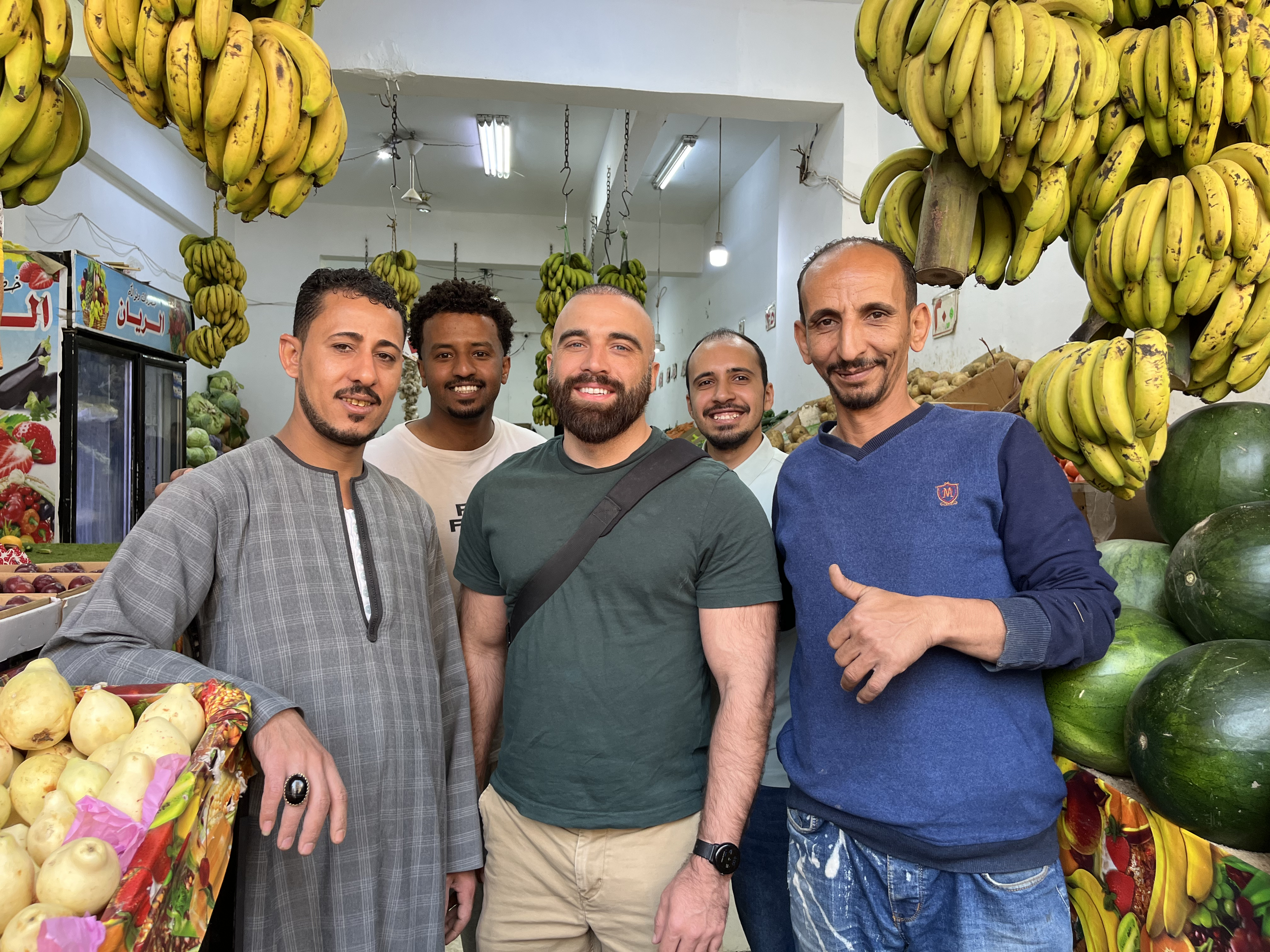 Group of five men smiling at camera inside a fruit market, with numerous bunches of bananas hanging overhead and various fruits on display.