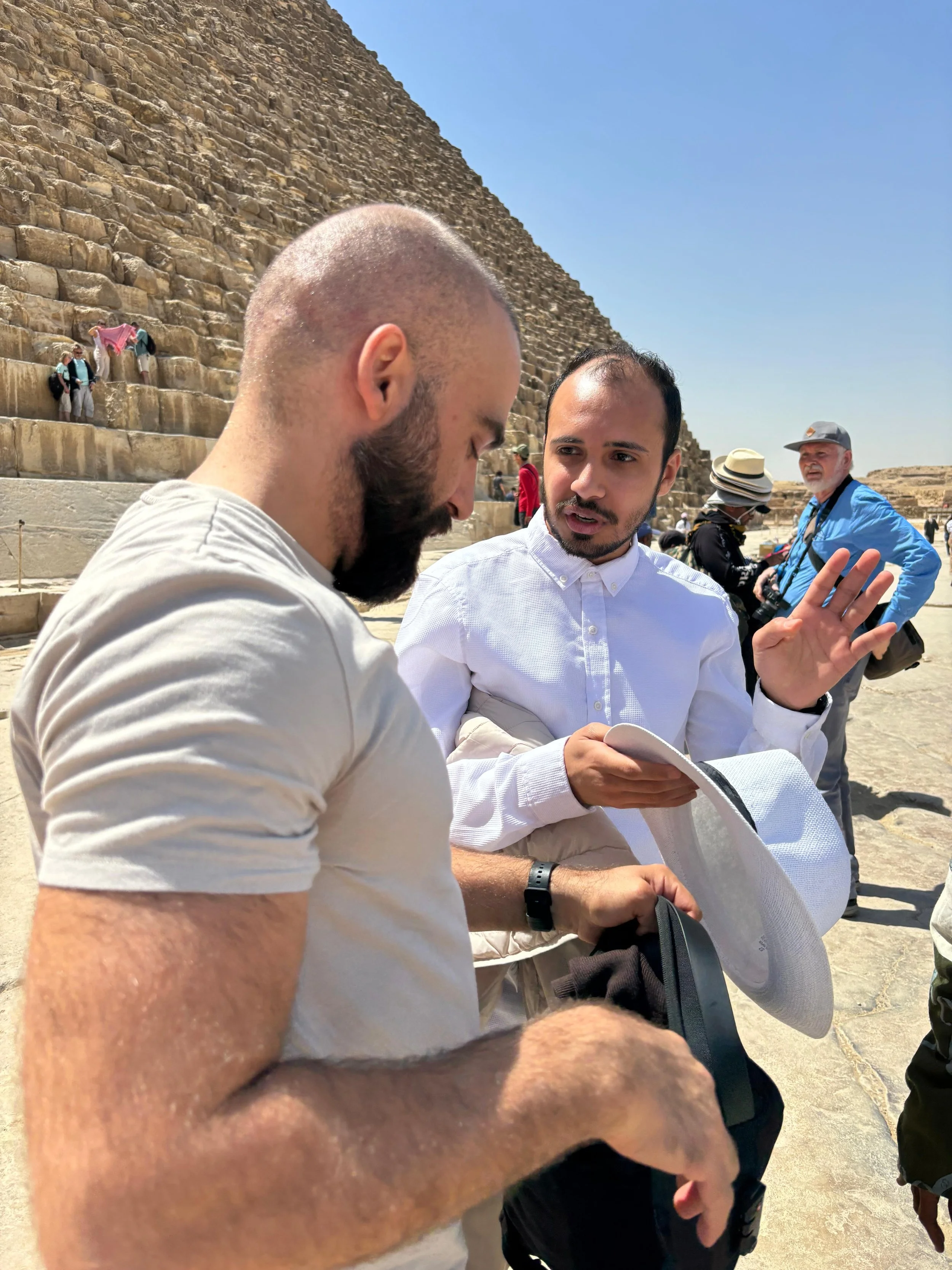 Two men are engaged in a conversation near the pyramids in Egypt. One man with a beard and shaved head is in the foreground, and another man with short dark hair is speaking. In the background, tourists, including a man with a camera and a woman with a hat, can be seen taking photos and exploring the site under a clear blue sky.