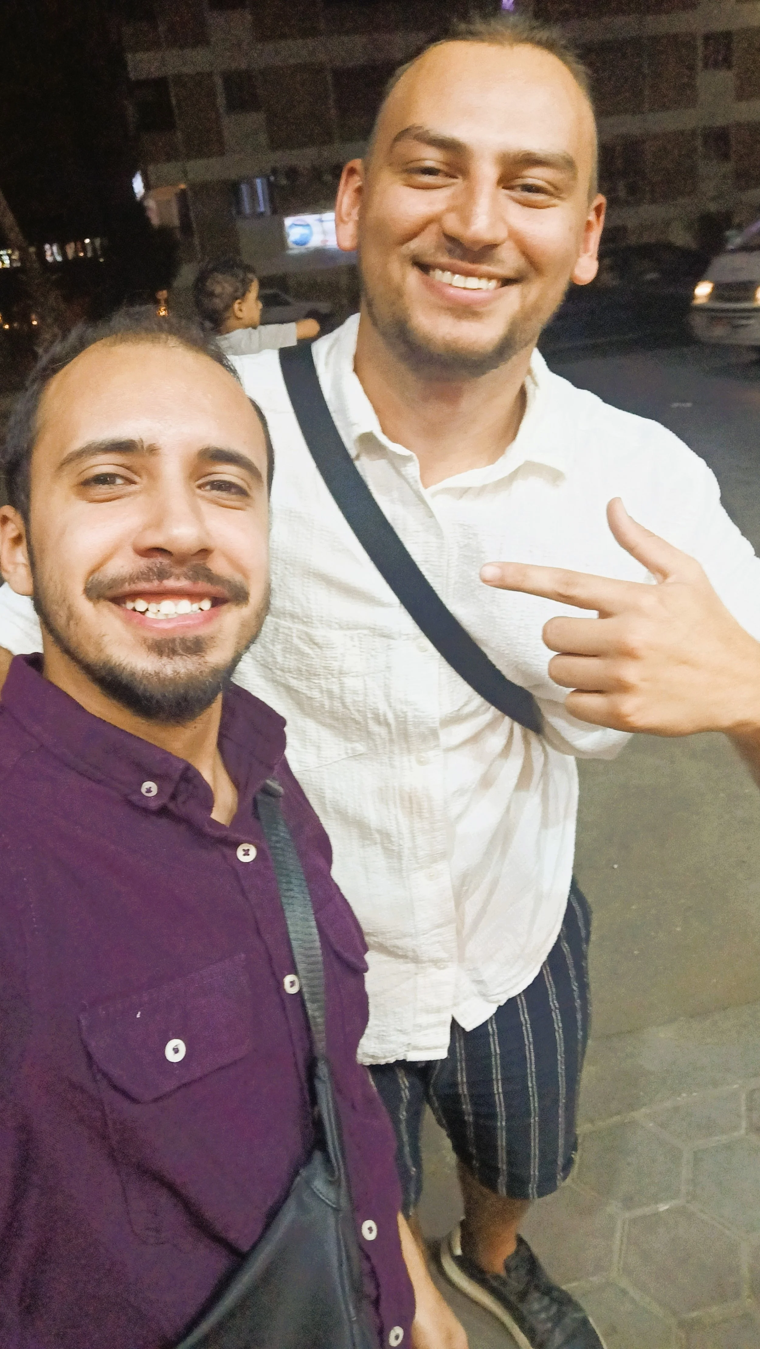 Two men smiling, taking a selfie at night. One man is pointing at the other. There are people in the background, and the scene appears to be outdoors near a building with parked cars.