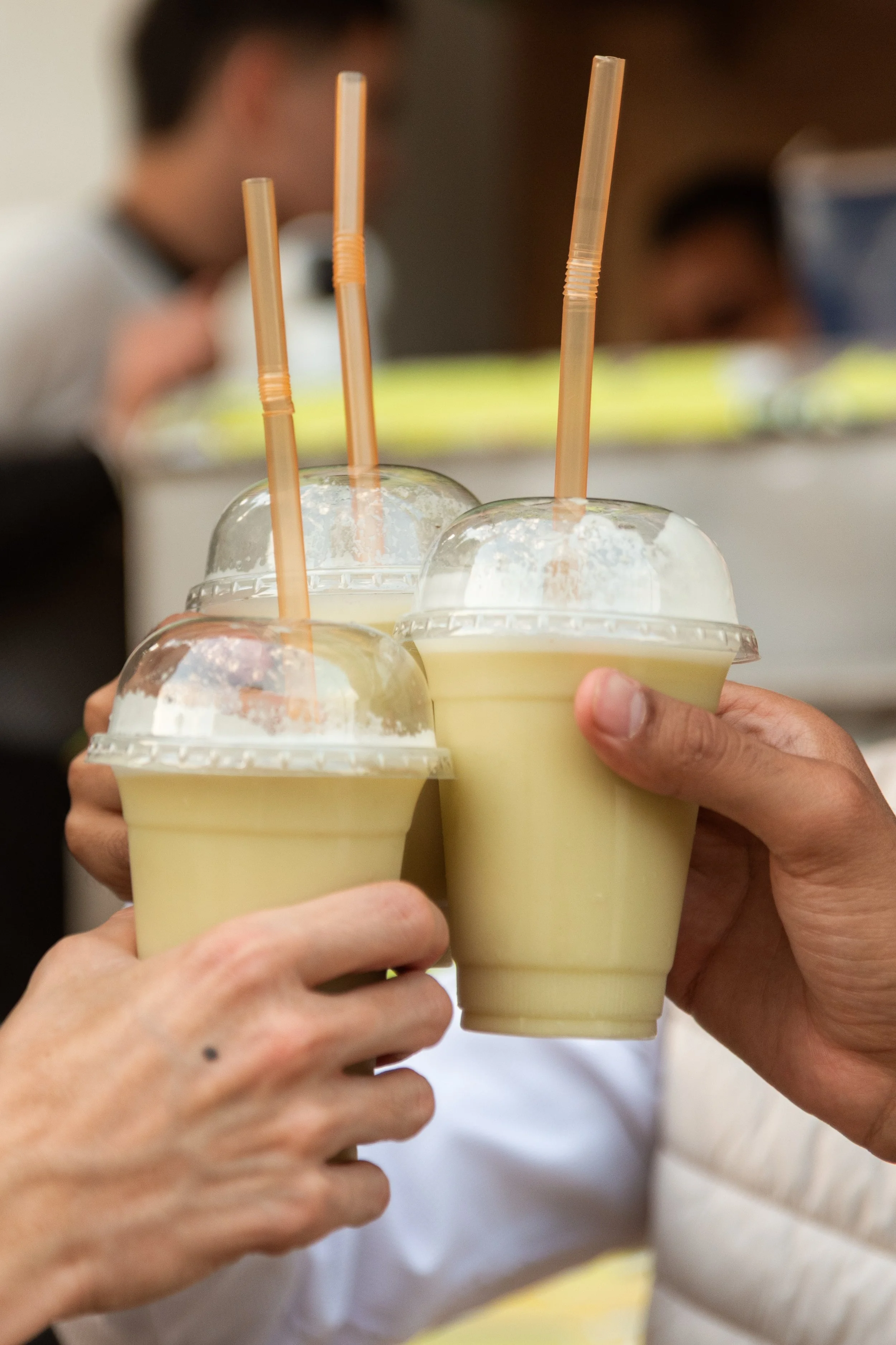 Multiple hands holding cups of a yellow-colored drink with dome-shaped plastic lids and orange straws, blurred background of people.
