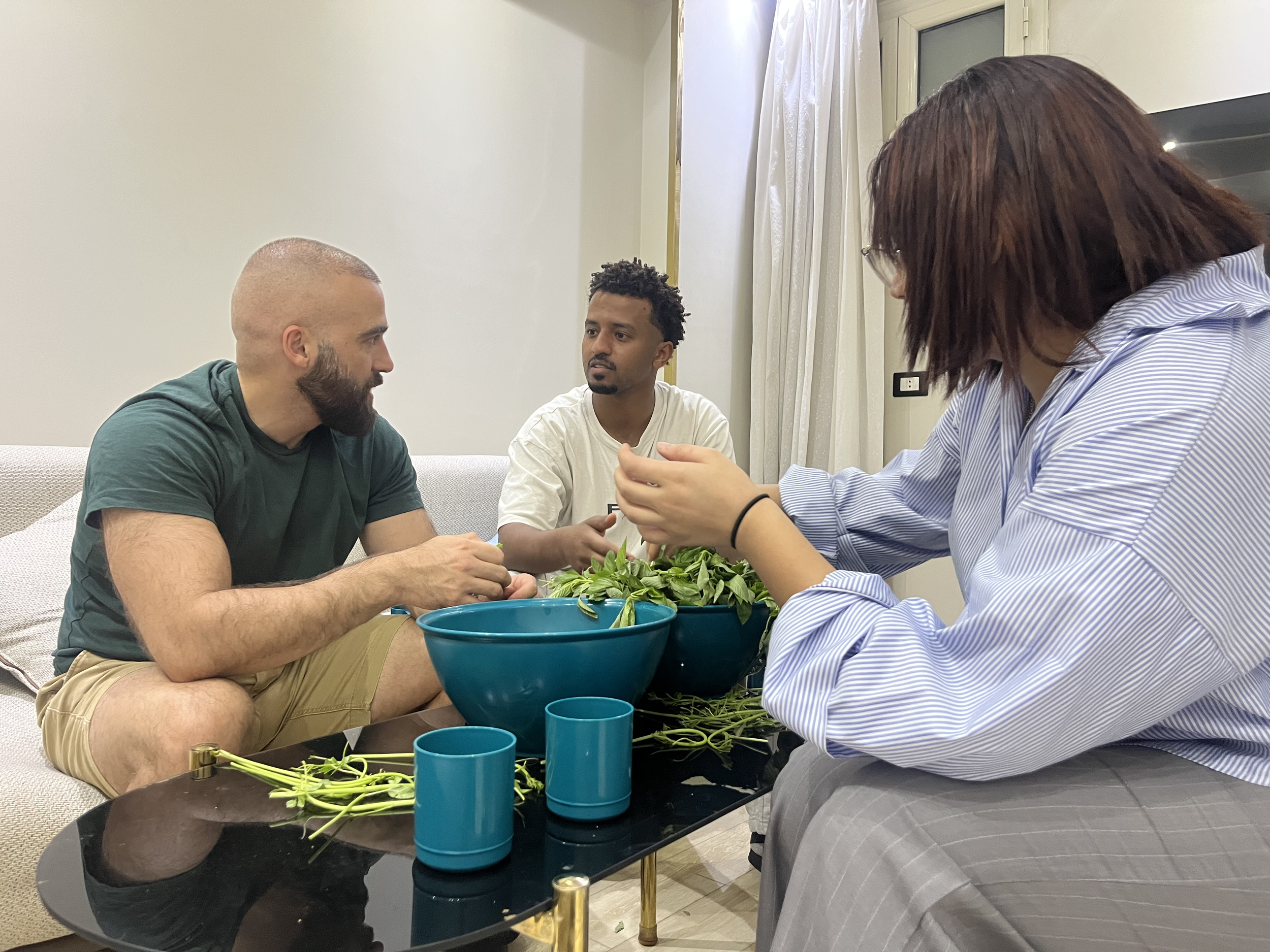 Three people sitting around a coffee table, making a craft with green plant stems, with a large bowl and blue cups on the table. The scene is indoors with cream-colored walls and a window with curtains.