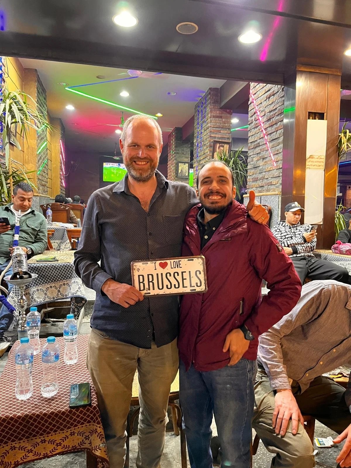 Two men smiling at a restaurant, holding a sign that reads "I love Brussels". One man is wearing a dark button-up shirt, and the other is wearing a red jacket. There are other diners and a TV screen in the background.