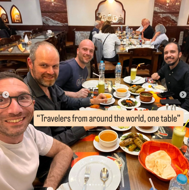 Four men sitting at a large table filled with bowls of soup, dishes, and drinks, in a restaurant with other diners in the background. They are smiling and appear to be enjoying a meal together.