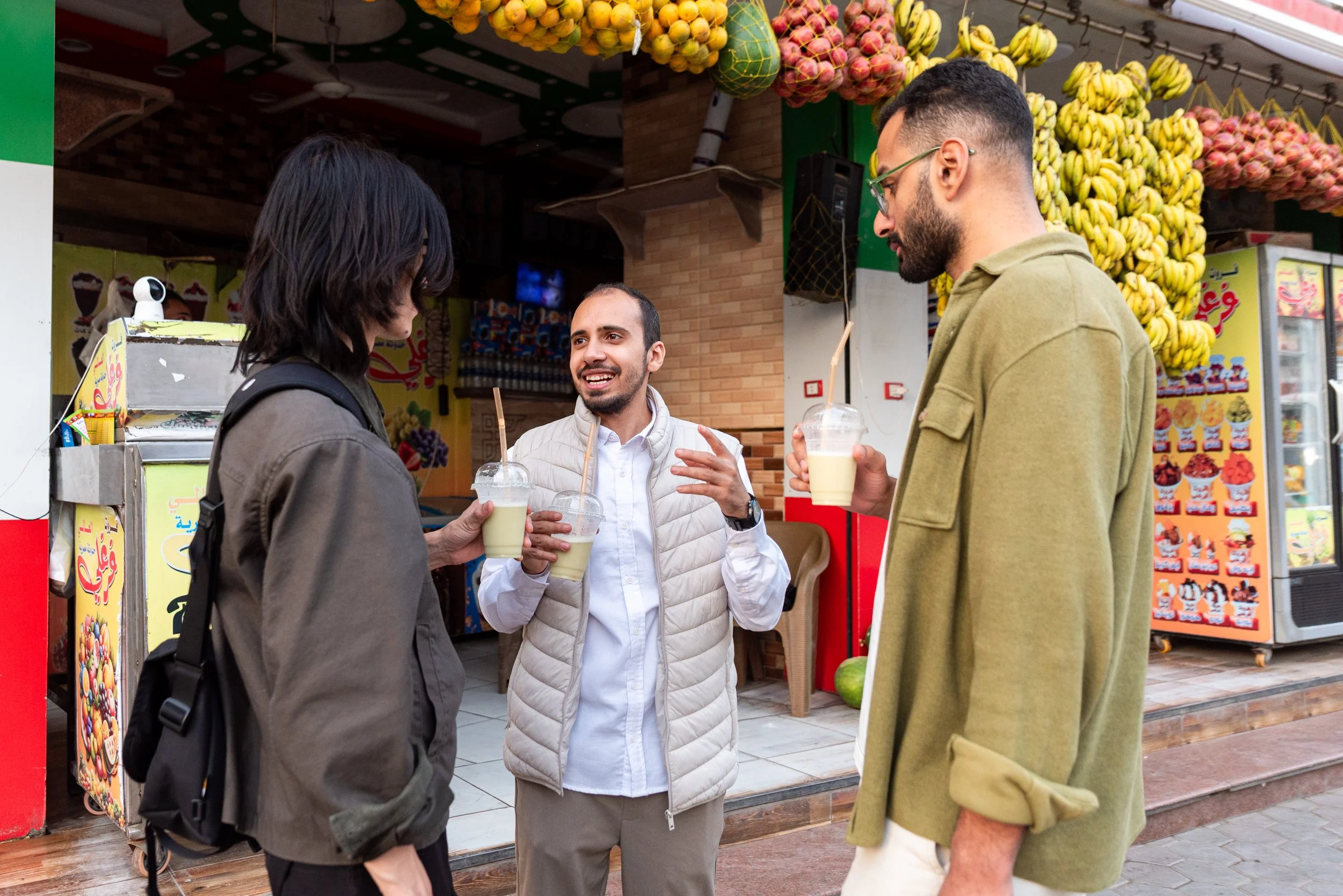 Three people standing outside a fruit stand, holding drinks with straws, engaged in conversation. There are bananas and other fruits hanging overhead in the background.