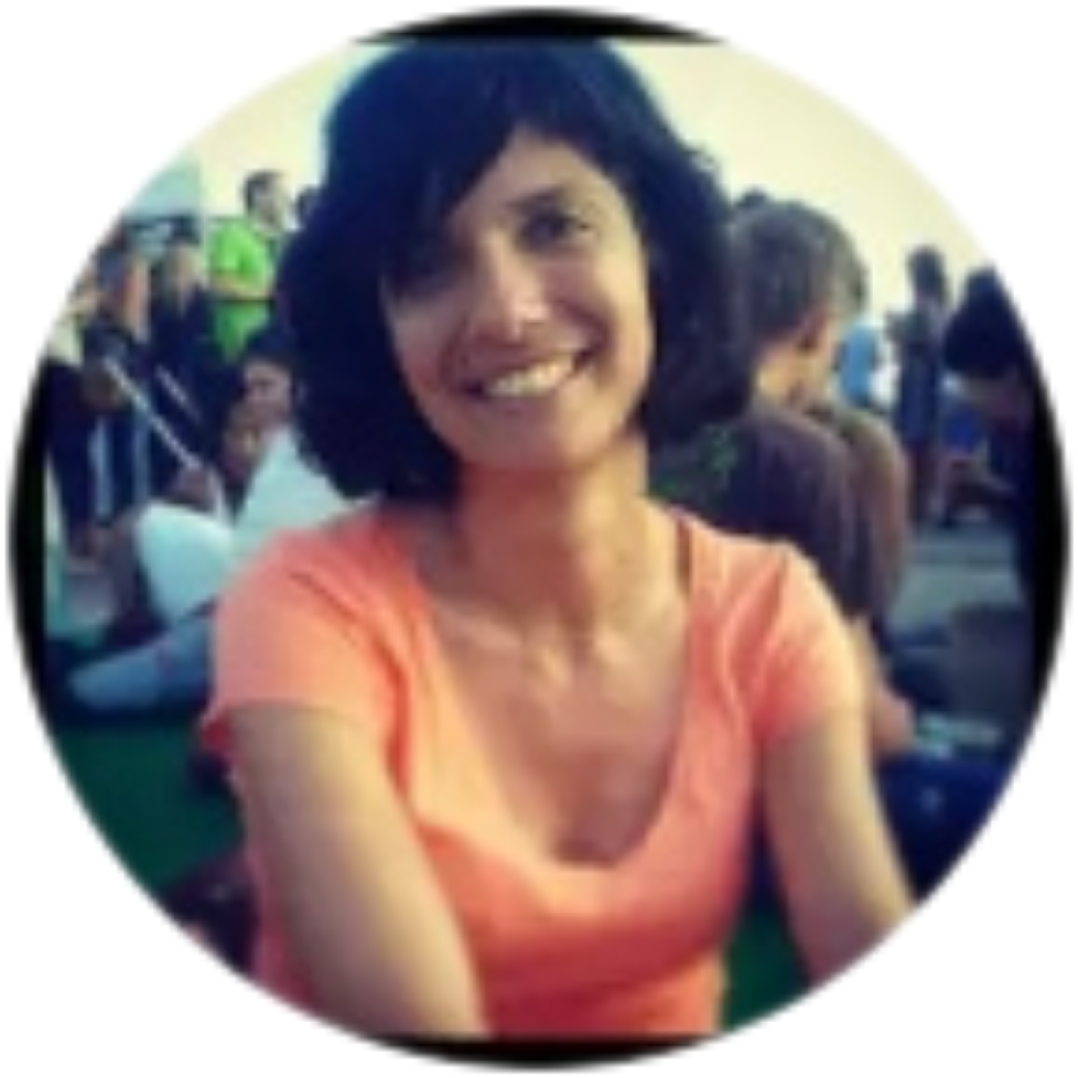 Smiling woman with short dark hair wearing an orange top, sitting outdoors at a social gathering with other people in the background.
