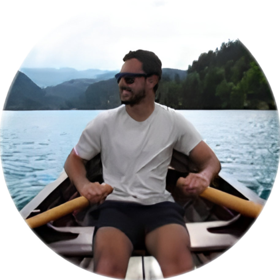 Man wearing sunglasses, white t-shirt, and black shorts, smiling while rowing a boat on a lake with mountains and trees in the background.