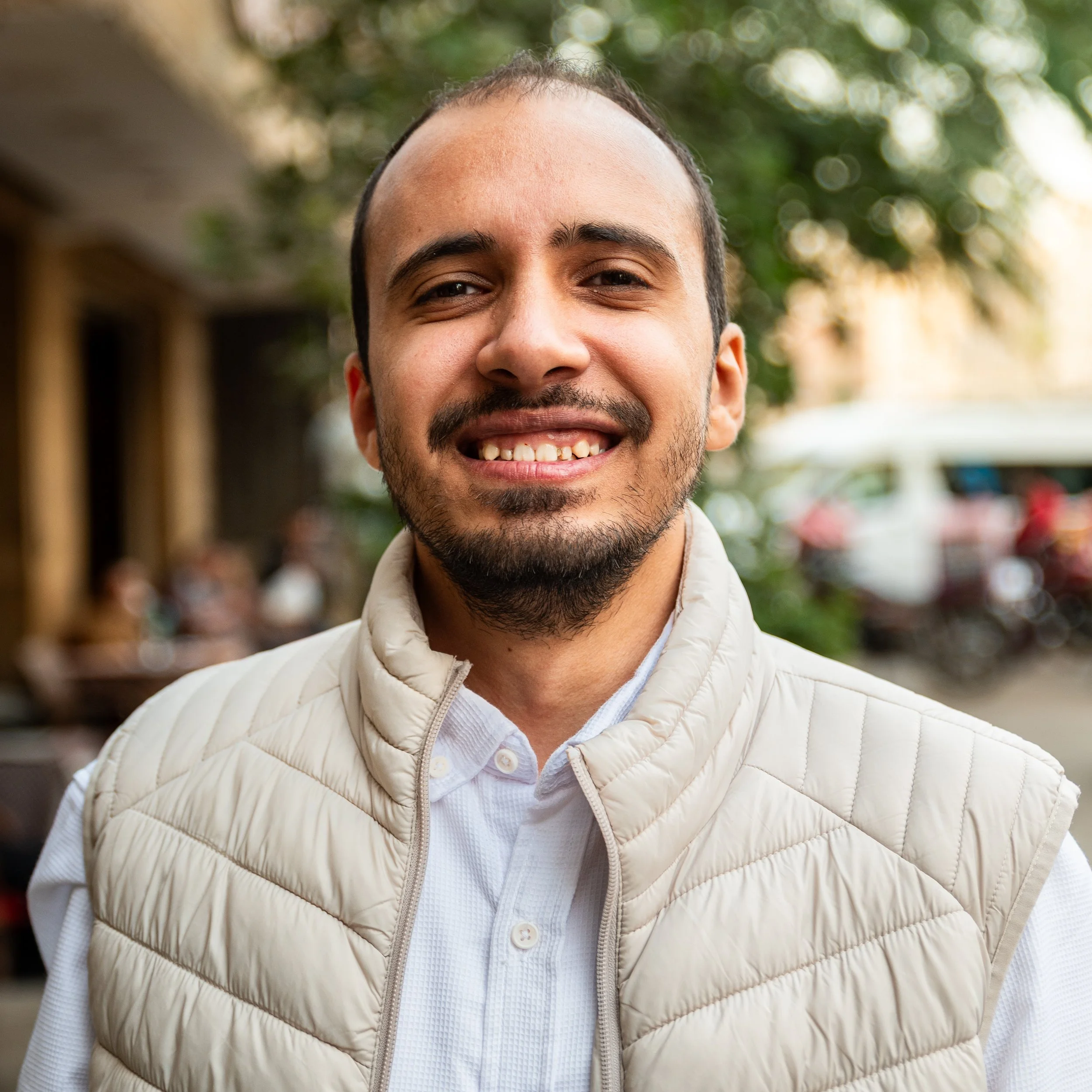 A close-up of a smiling man with a beard and mustache, wearing a white quilted jacket and a collared shirt, standing outdoors with trees and a building in the background.