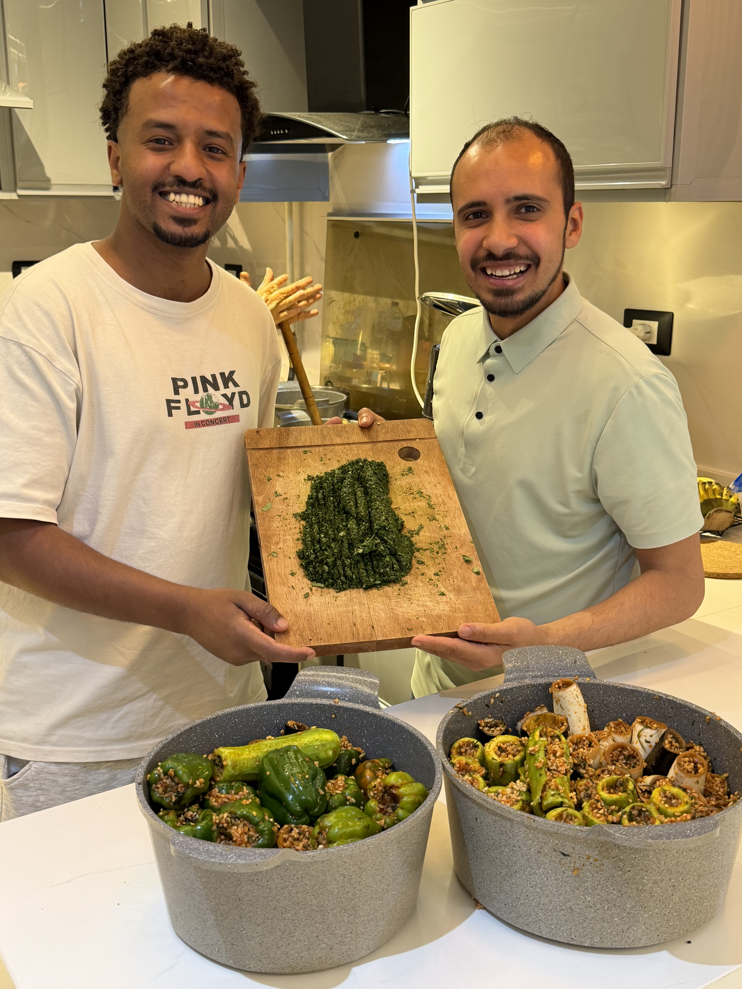 Two men smiling and holding a wooden cutting board with cooked or prepared kale in a kitchen. The front of the image shows two large bowls of green peppers filled with a mixture of grains and seasonings. The men are standing behind a counter with cooking utensils and food in a home kitchen setting.