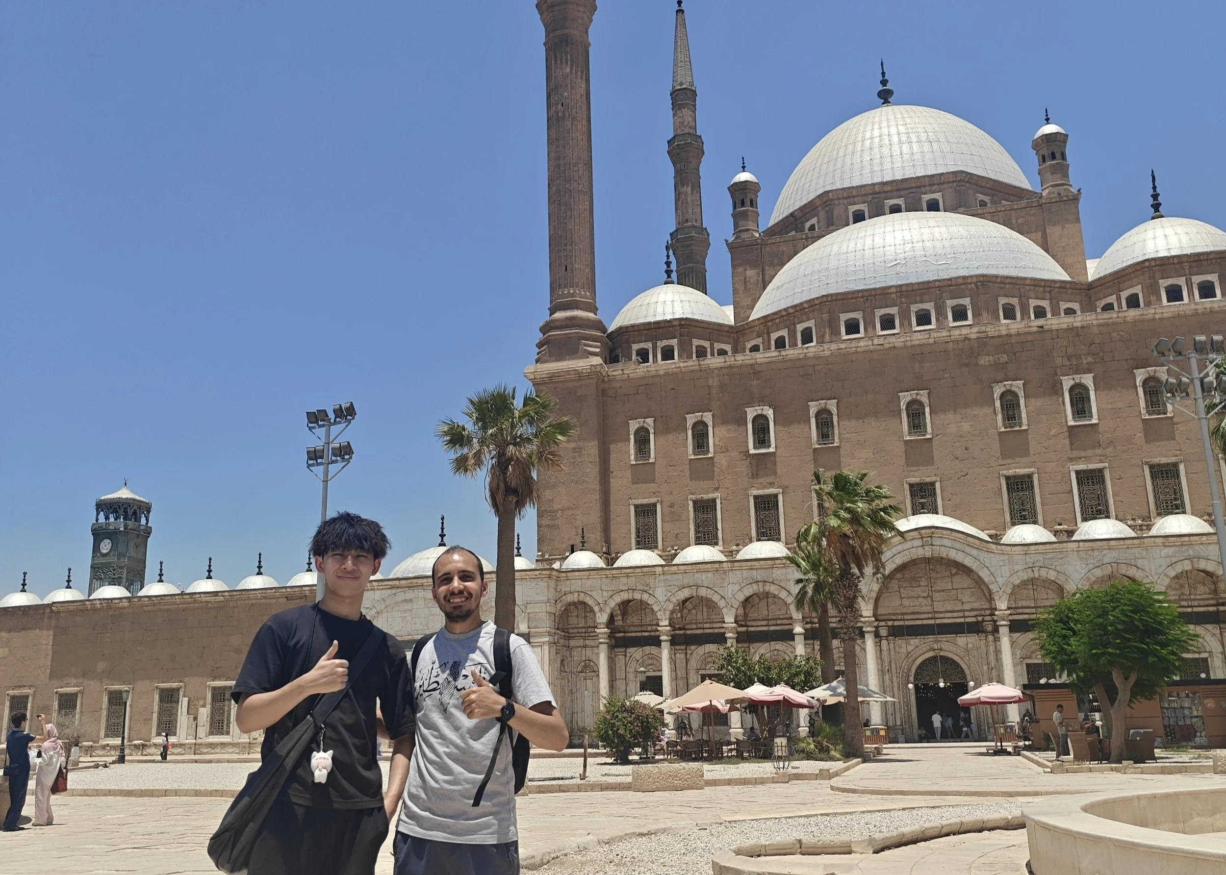 Two men standing in front of a historic building with domes and minarets, under a clear blue sky, with a clock tower and palm trees nearby.